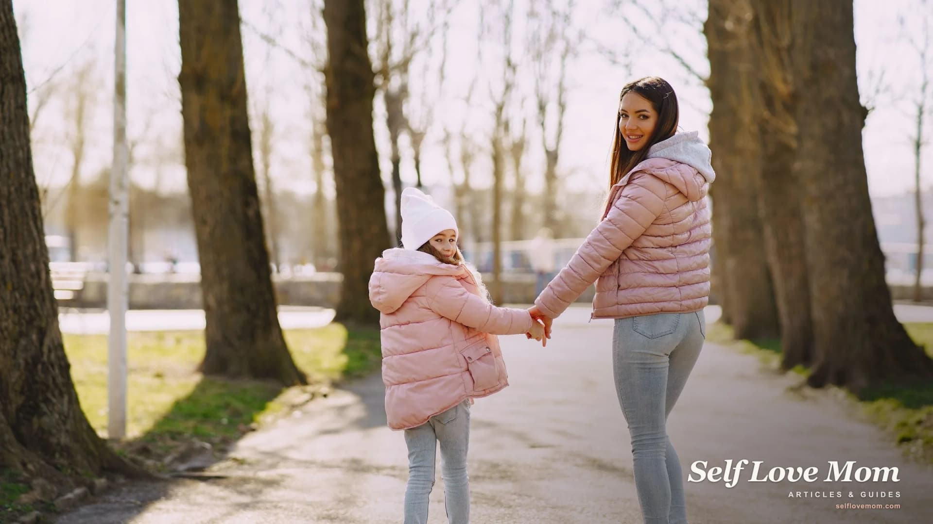 A mother and her young daughter hold hands on a sunlit park path, the calm a clear co-parent agreement helps protect.