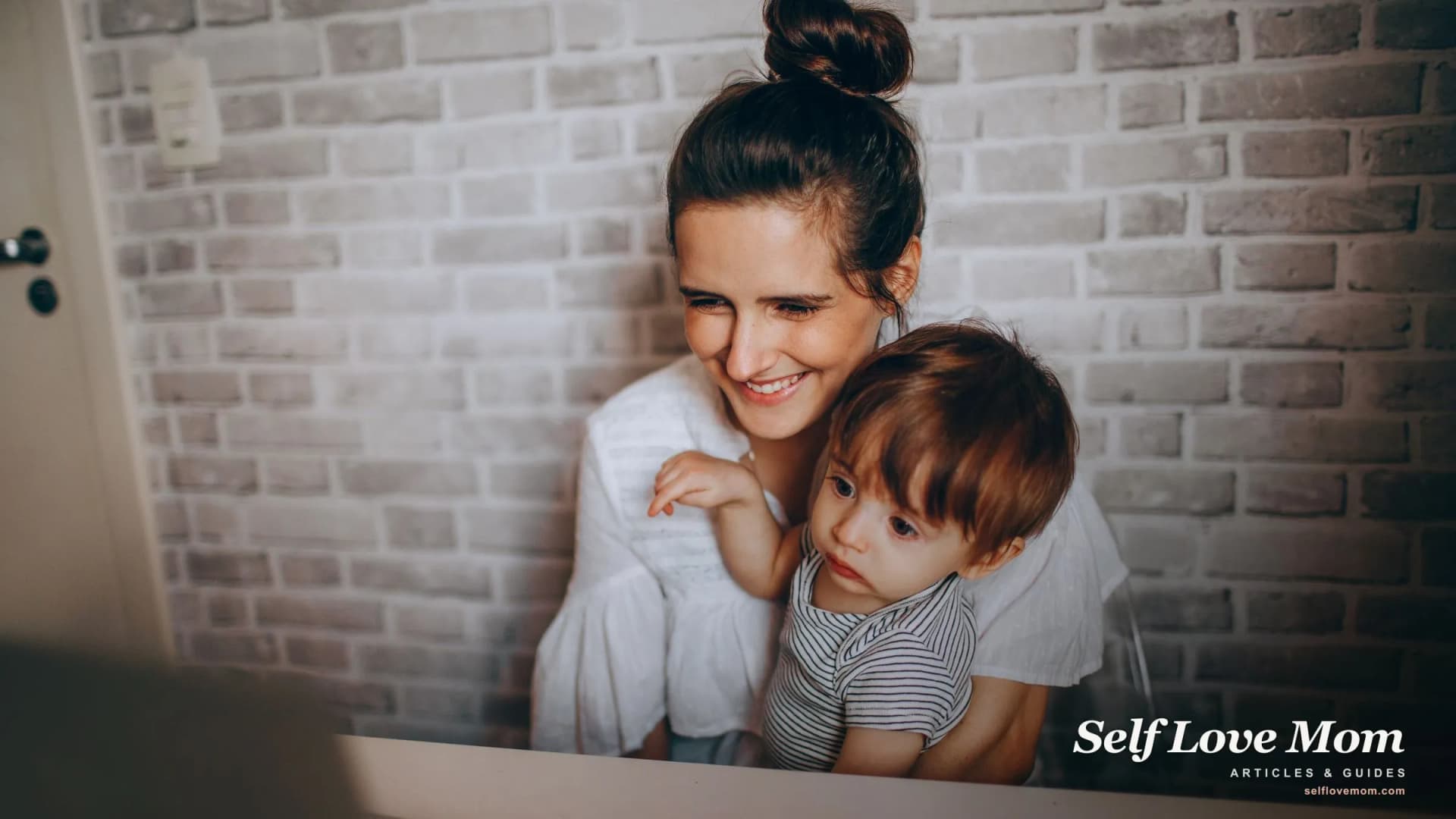 A single mom smiles while holding her toddler at her laptop, working from home with no second adult to help.