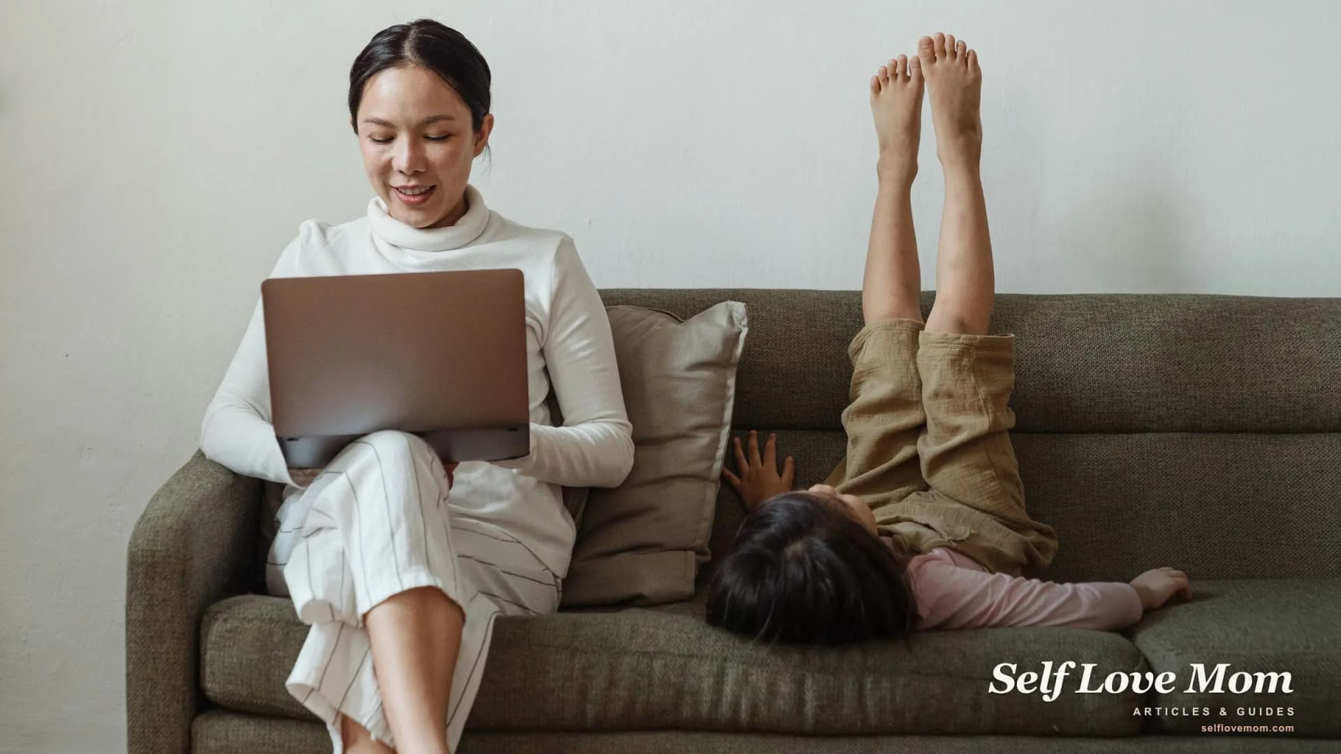 A working single mom plans her day on her laptop while her daughter relaxes beside her on the couch.