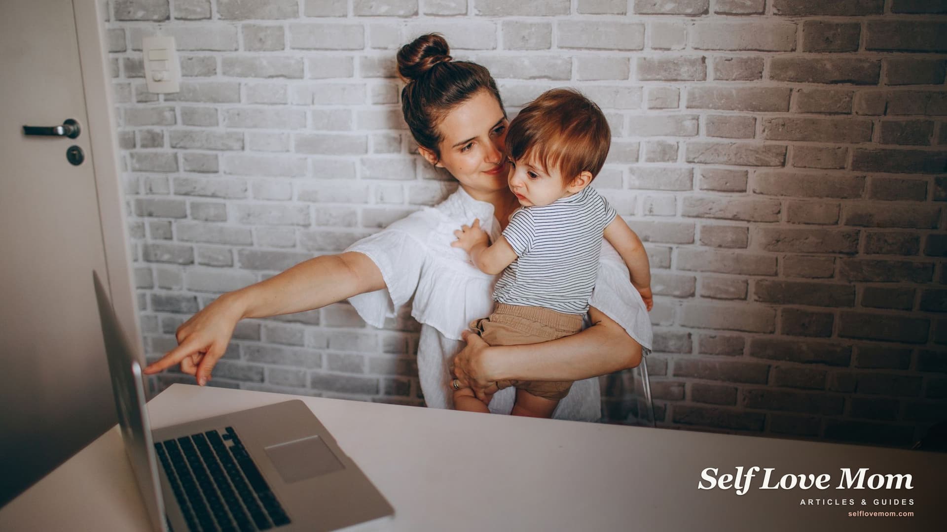 A single mother works on her laptop while holding her toddler at home, balancing a flexible work-from-home job with childcare.