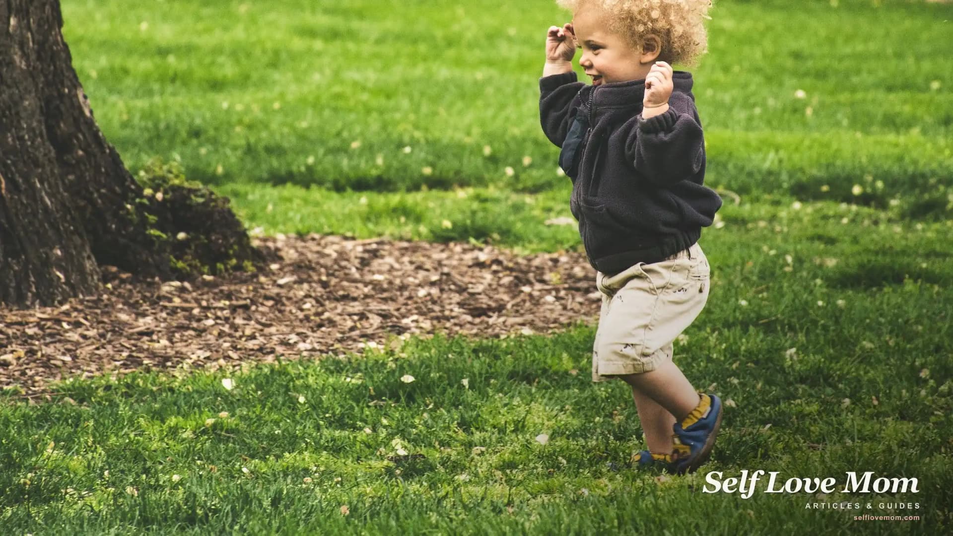 A curly-haired toddler runs across the grass in a park, mid-stride and grinning.