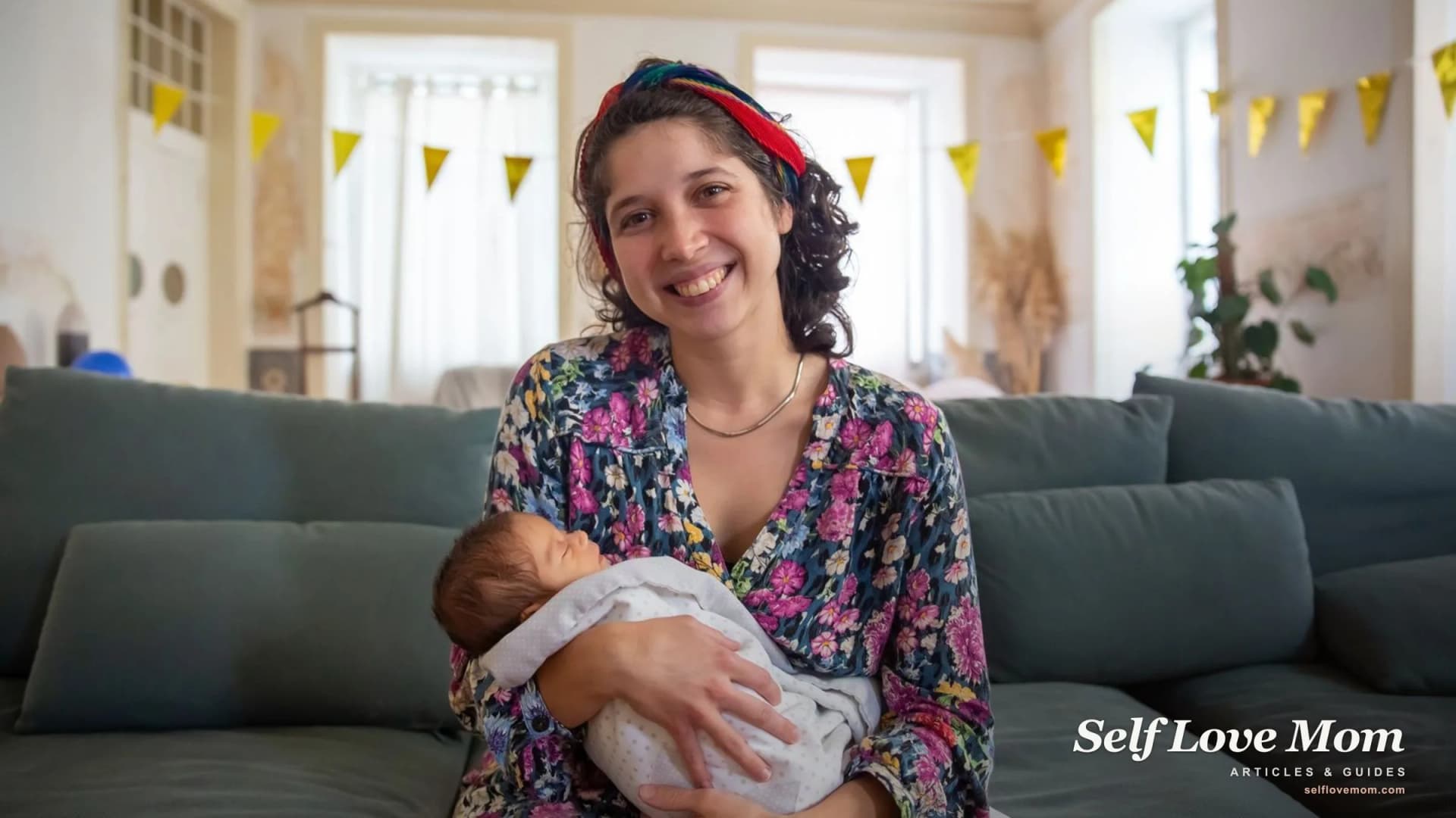 A smiling new mother sits on the sofa cradling her sleeping newborn.