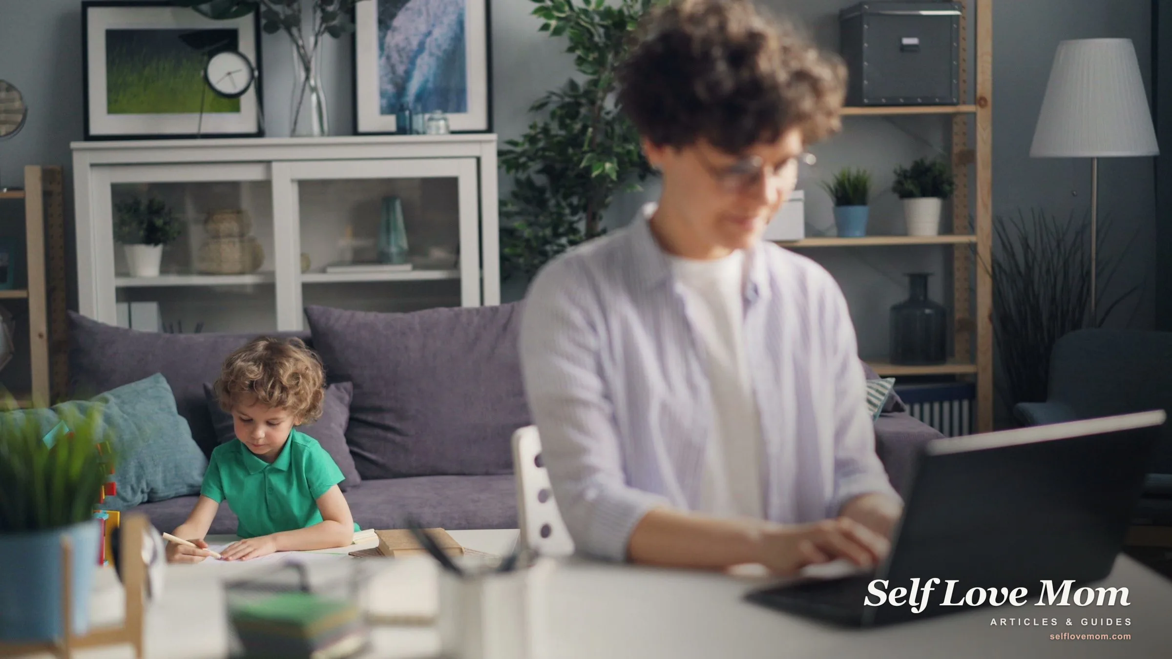 A single mom studies on her laptop at home while her young son draws nearby, fitting free GED classes around family life.