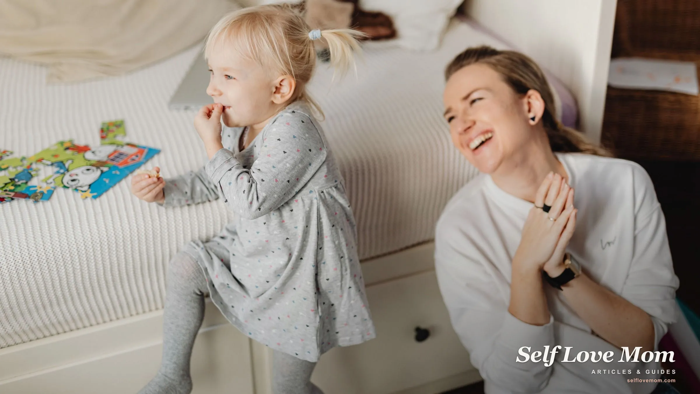 A stay-at-home mom laughs with her young daughter beside a puzzle on the bed during an everyday moment at home.