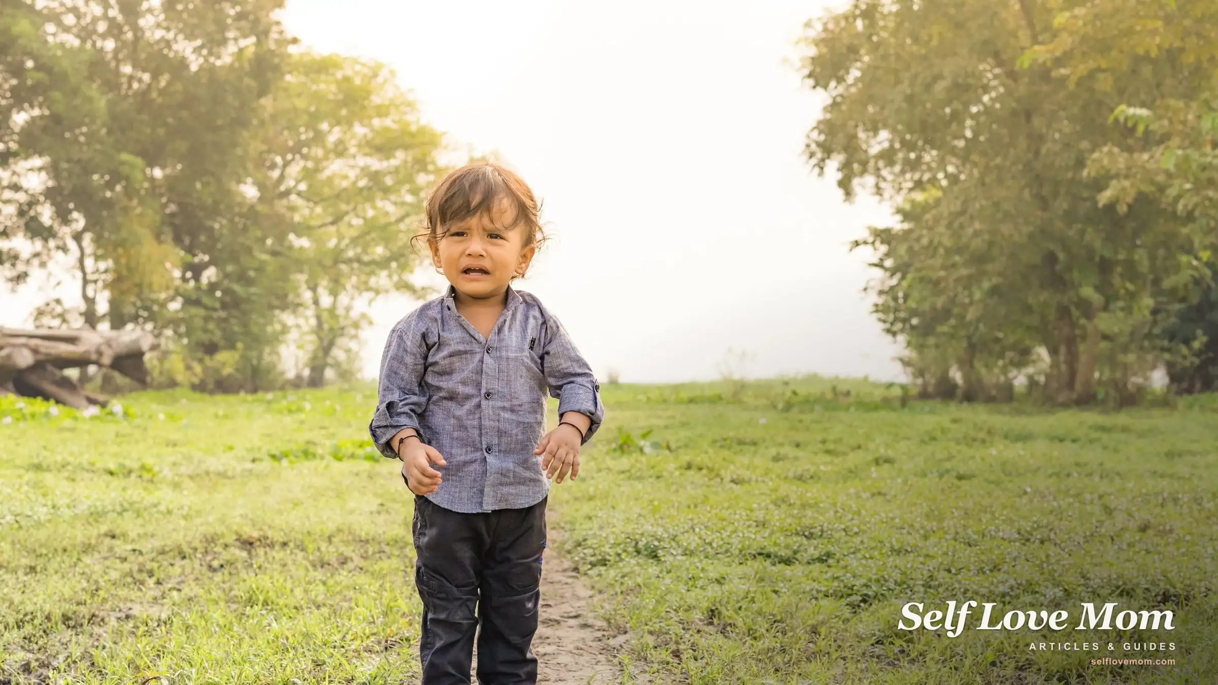 A toddler in a blue shirt stands smiling in a sunlit grassy field.