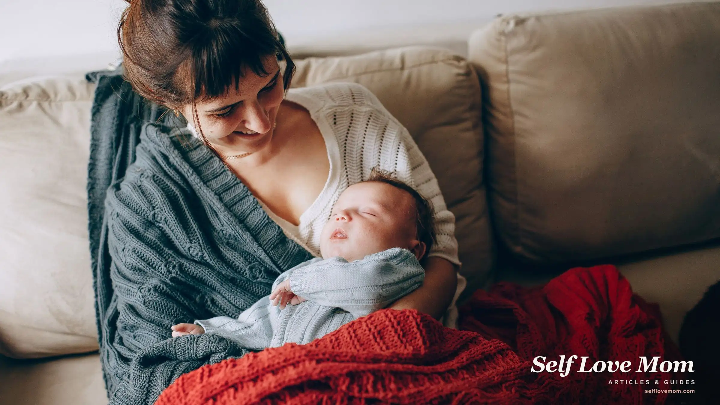 A smiling new mom cradles her sleeping newborn on the sofa, the calm a good self-care gift is meant to protect.
