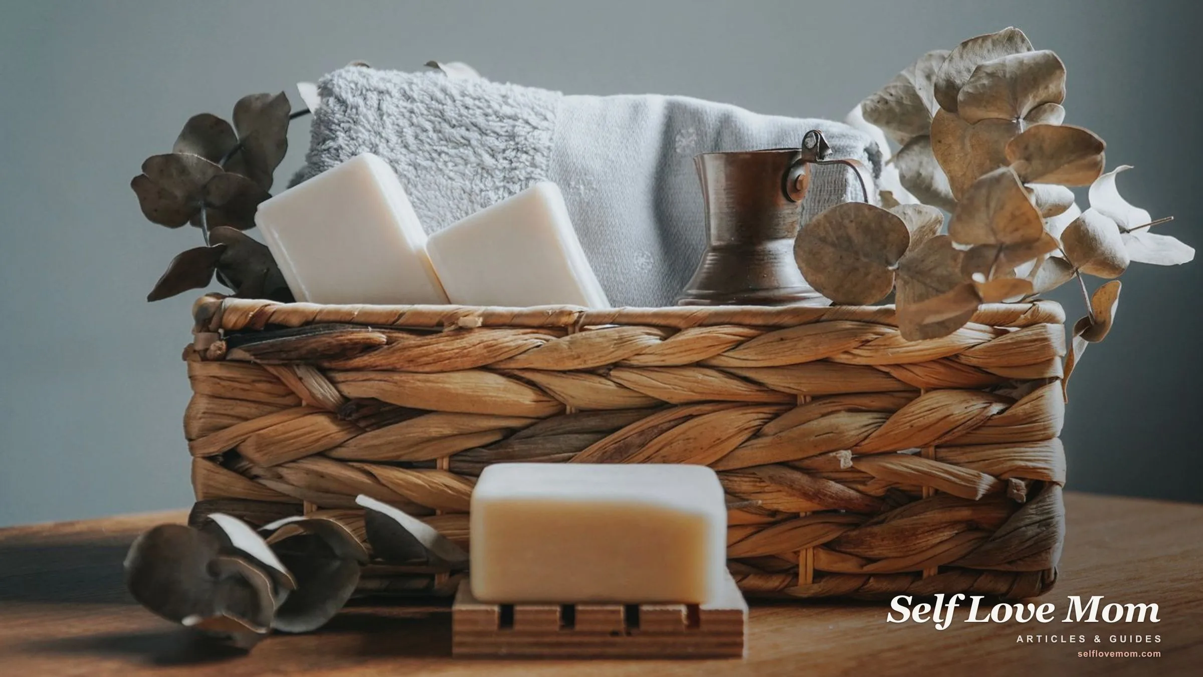A woven self-care basket filled with a folded towel, handmade soap, and dried eucalyptus on a wooden table.