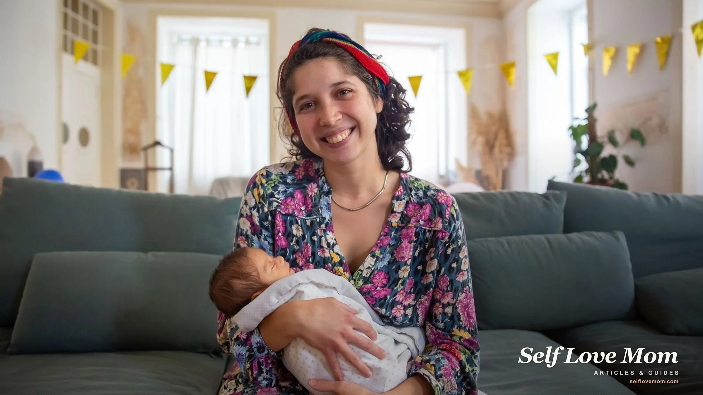 A smiling new mother sits on the sofa cradling her sleeping newborn.