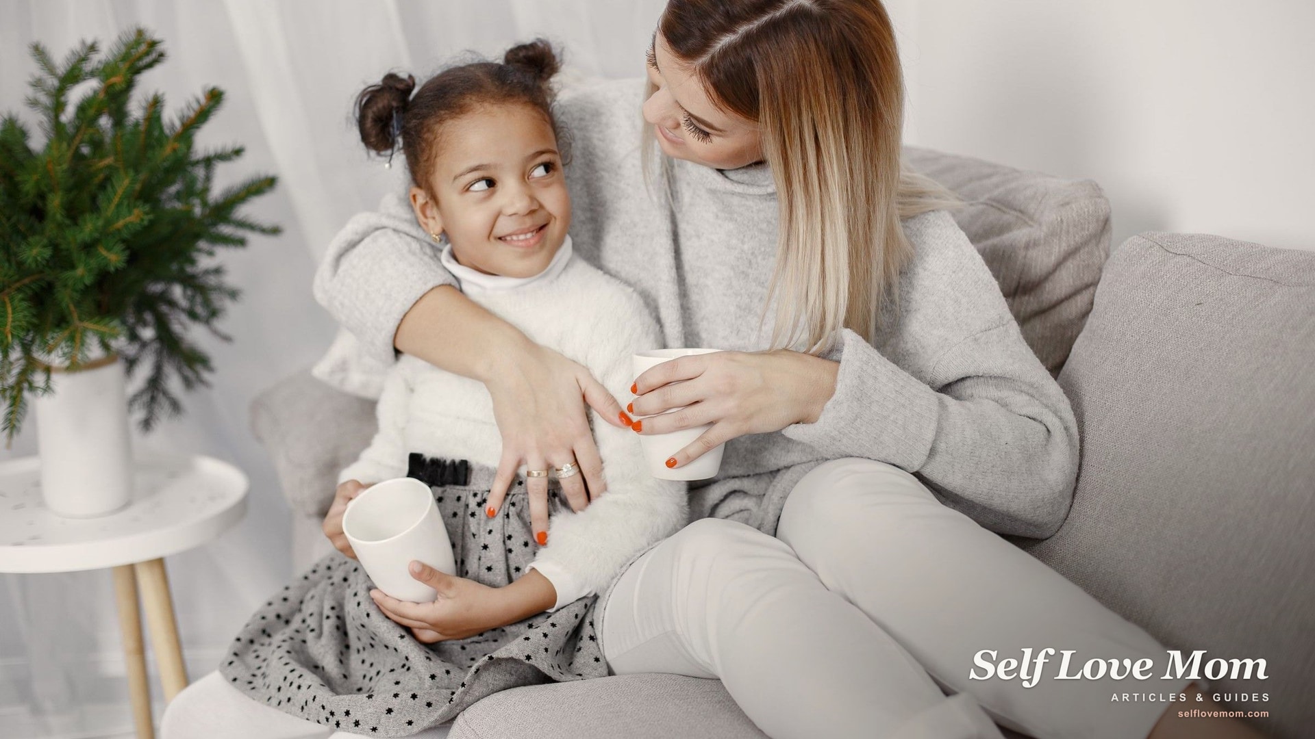 A mother and her young daughter share a calm moment over warm mugs on the couch, an everyday act of self care for mothers.
