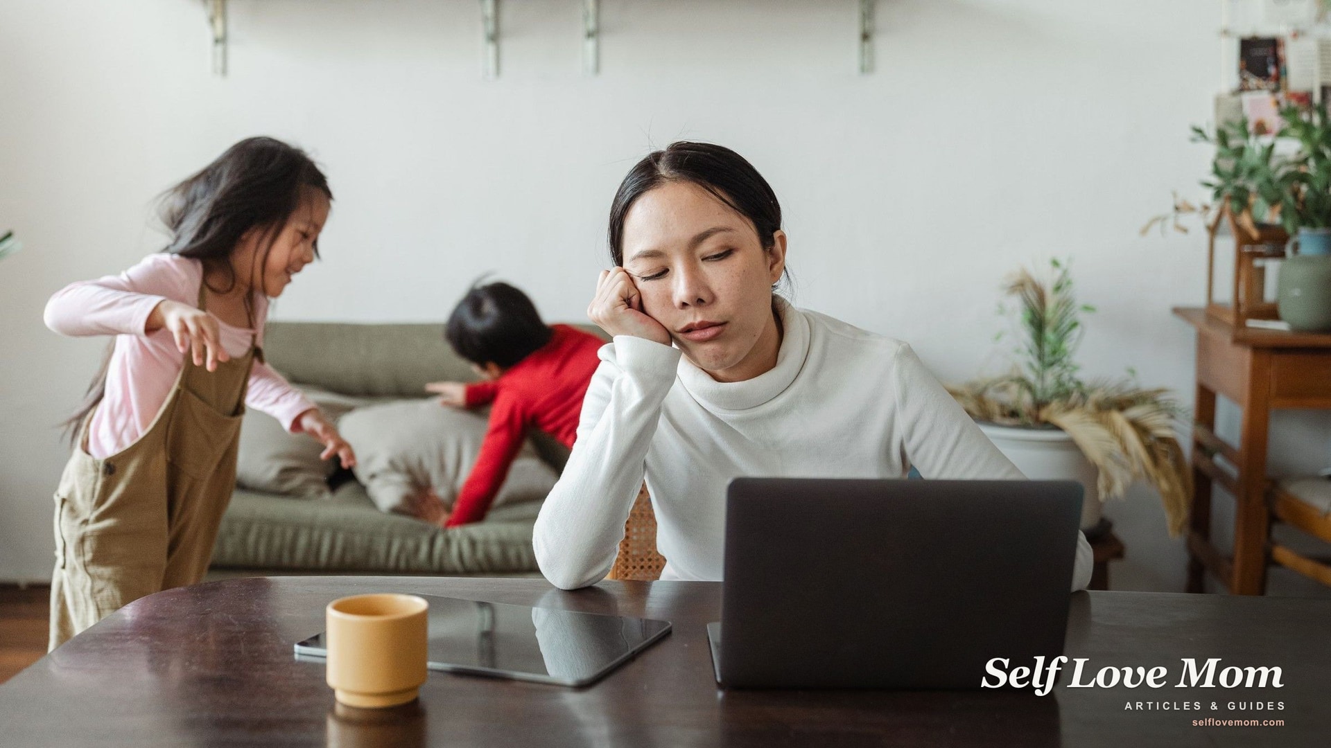 A tired mom leans on her hand at her laptop as her kids play behind her, the daily overwhelm behind mom burnout.