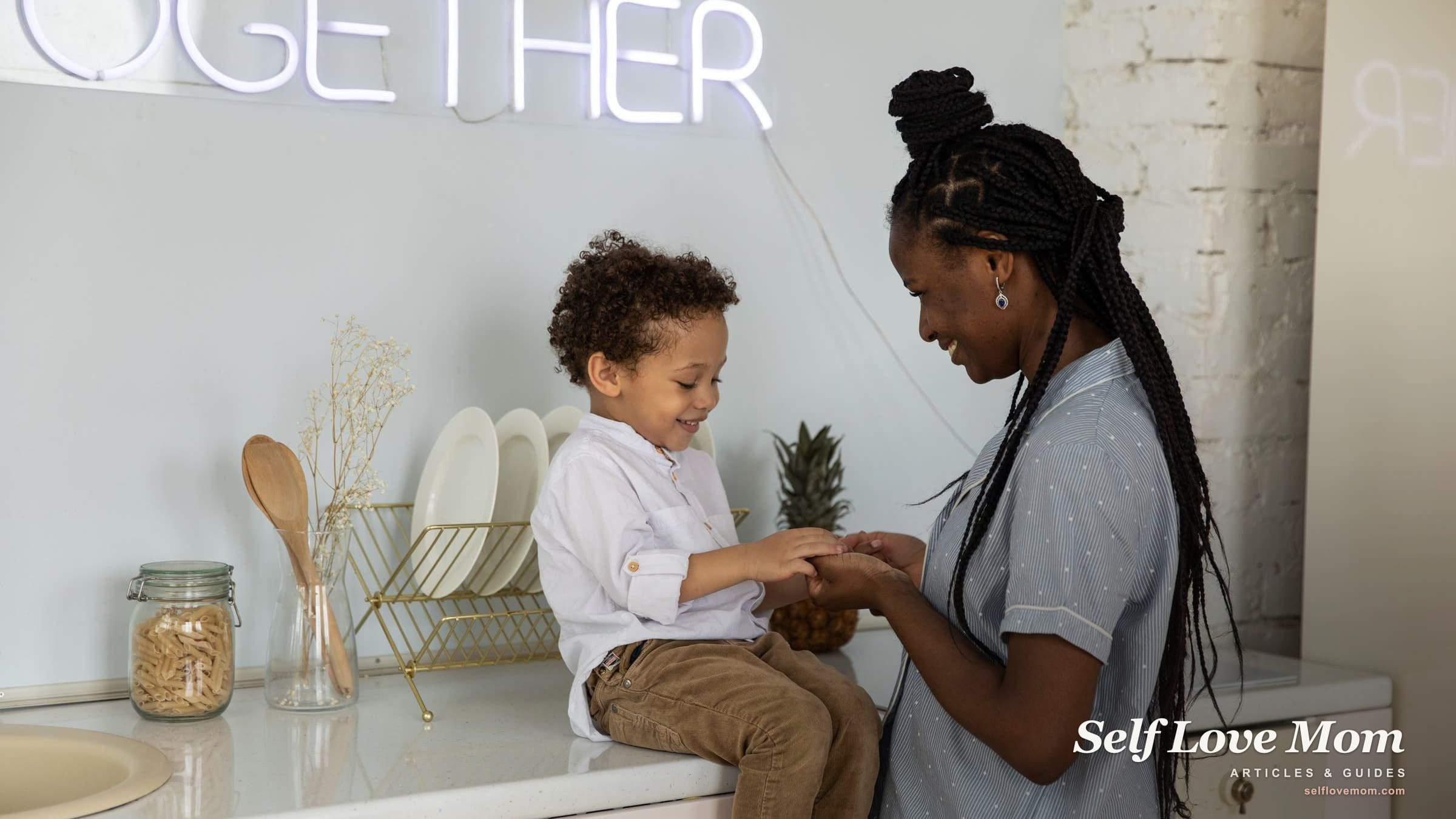 A mother holds her young son's hands while he sits on the kitchen counter beside the dish rack, both of them smiling.