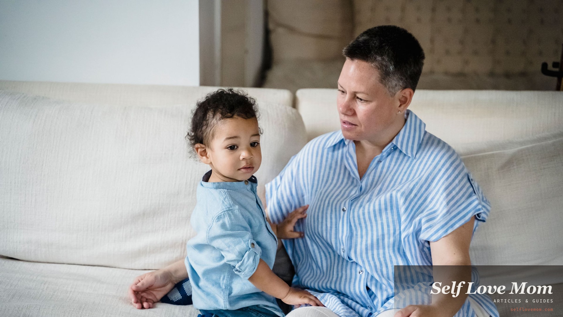 A mother sits on a sofa with her young son beside her, her attention somewhere else while he looks up at her