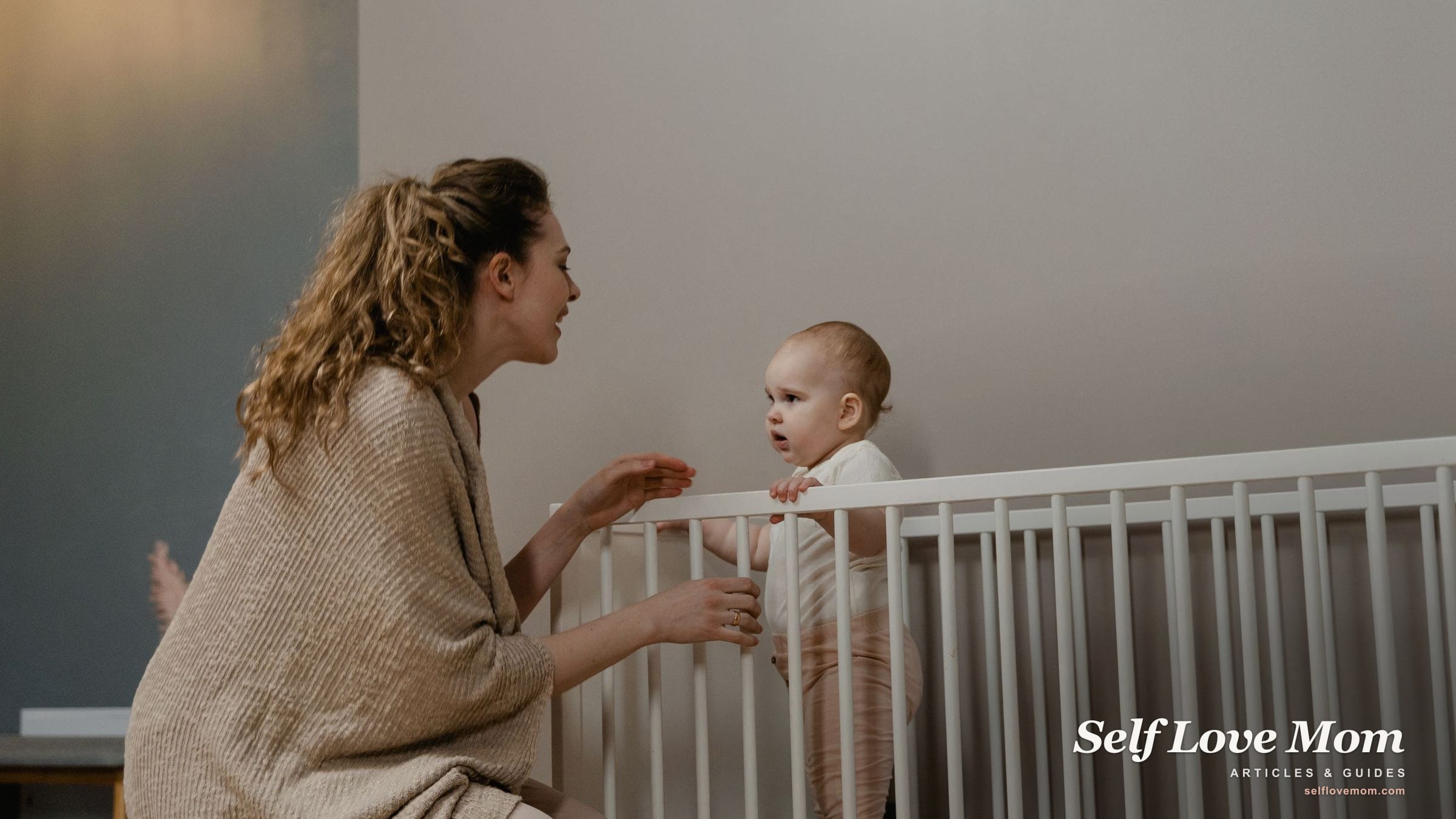 A smiling mother talks with her baby, who stands holding the rail of a bare white crib.