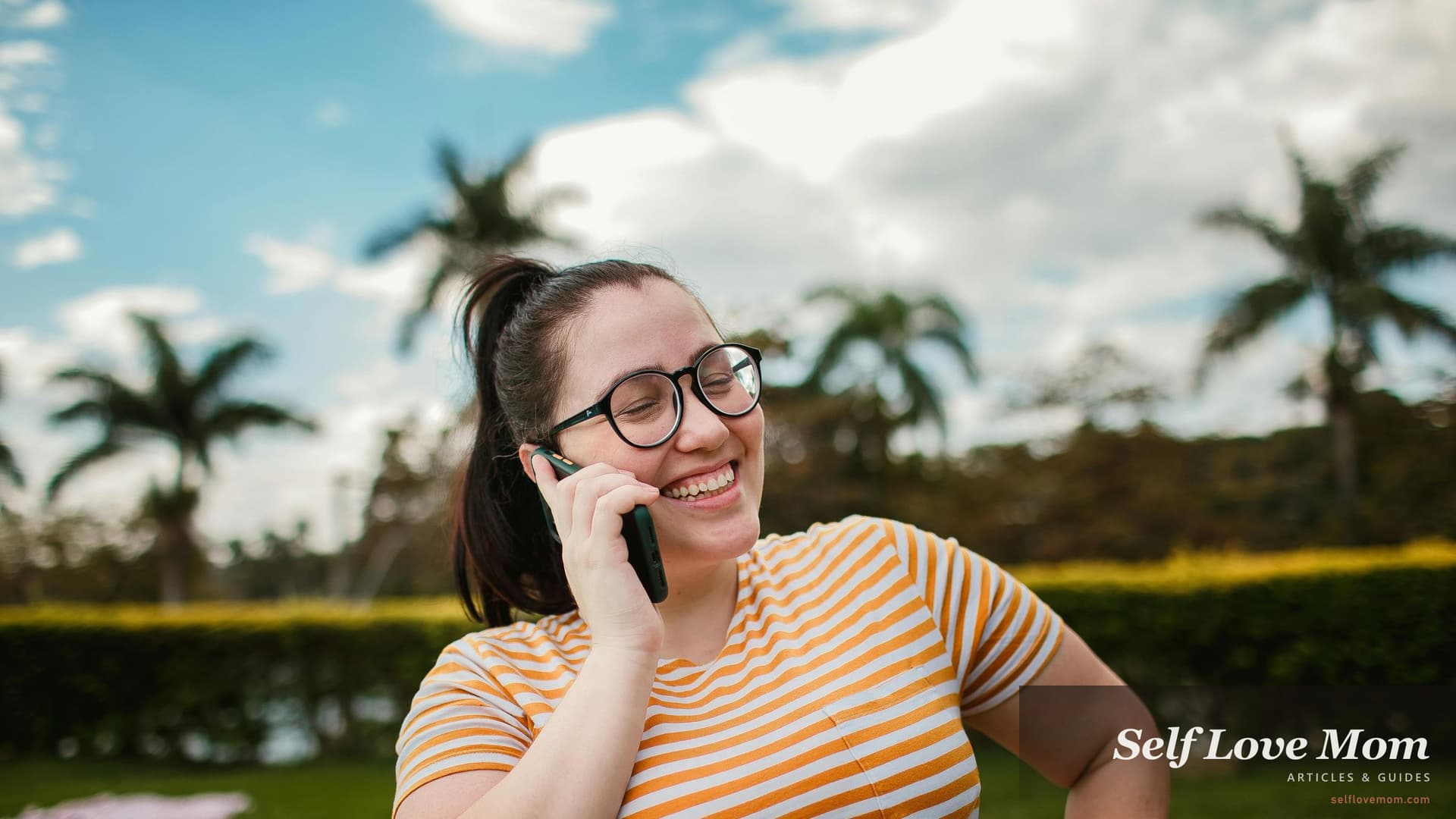 Single mom in a striped shirt smiling while talking on her phone outdoors with palm trees and bokeh in the background, the casual relaxed energy that the best dating sites for single moms are designed to support.