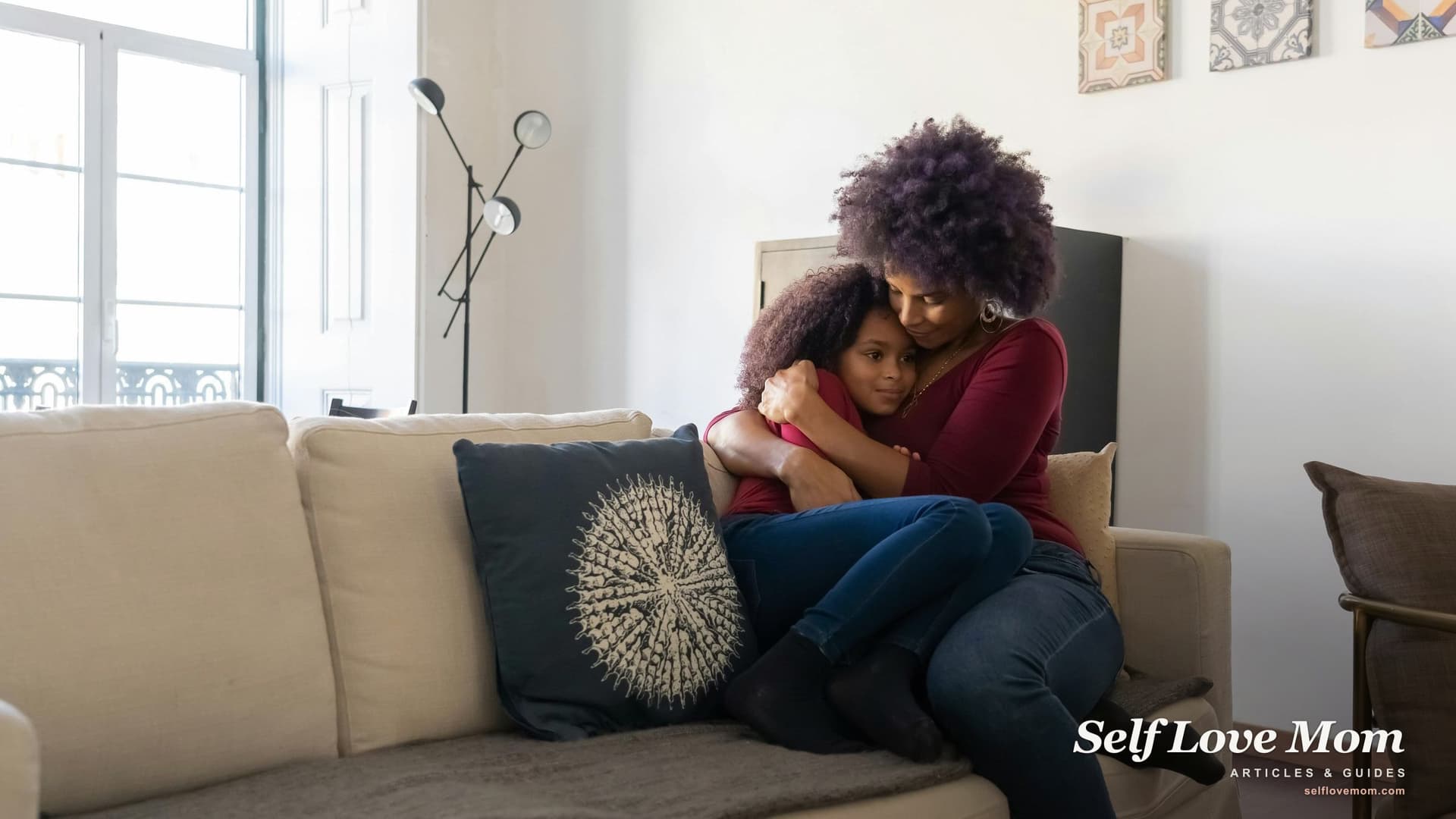 A mother sits on a cozy living-room sofa hugging her young daughter, both smiling in soft afternoon light.