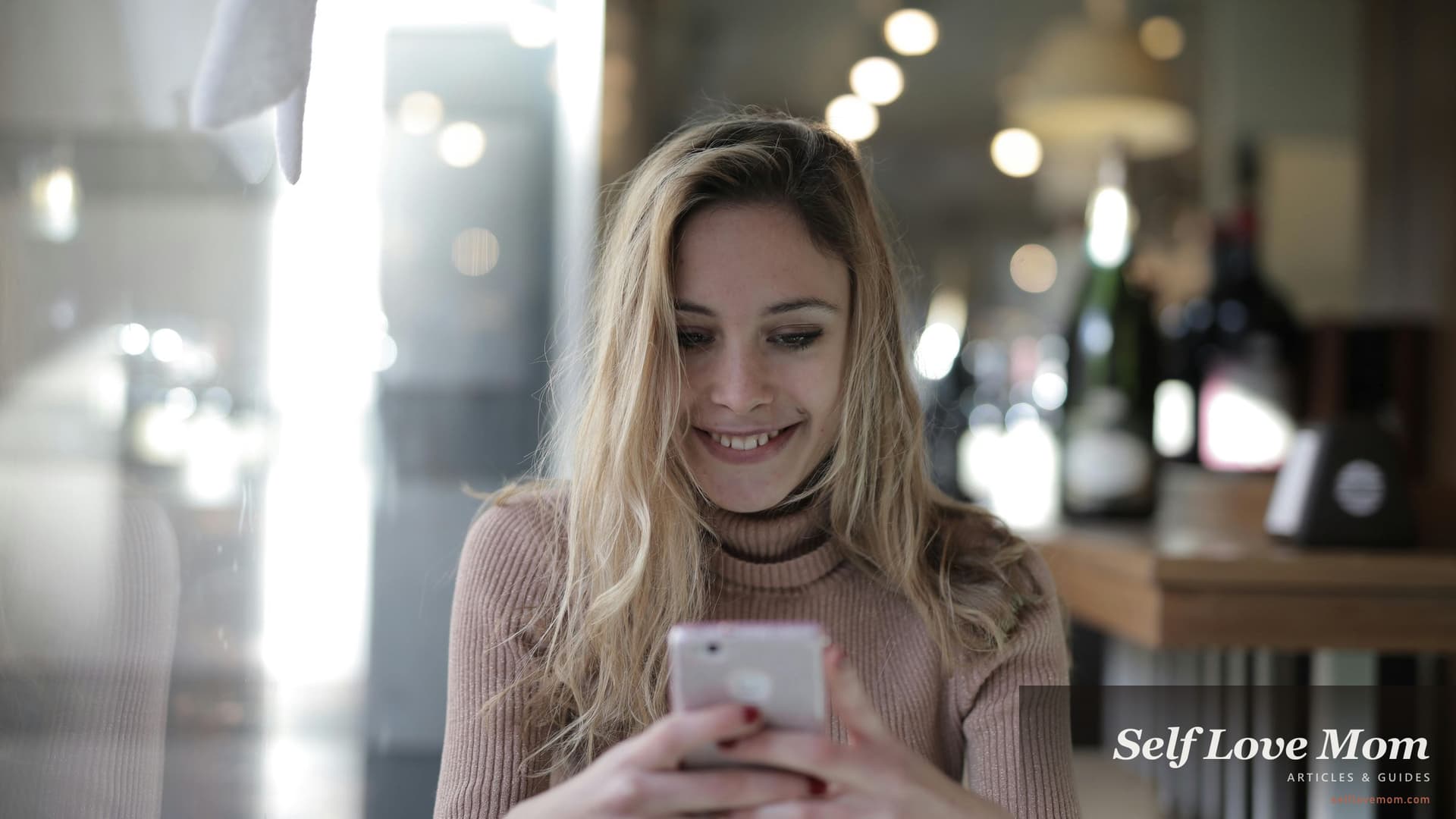 Single mom in a pink turtleneck smiling at her phone in a soft-lit cafe, the kind of relaxed everyday moment a strong dating profile is designed to invite into real life.