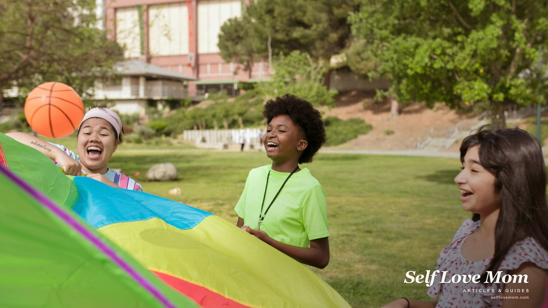 A group of children laugh and run through a colorful parachute on a sunny lawn at a summer day camp.
