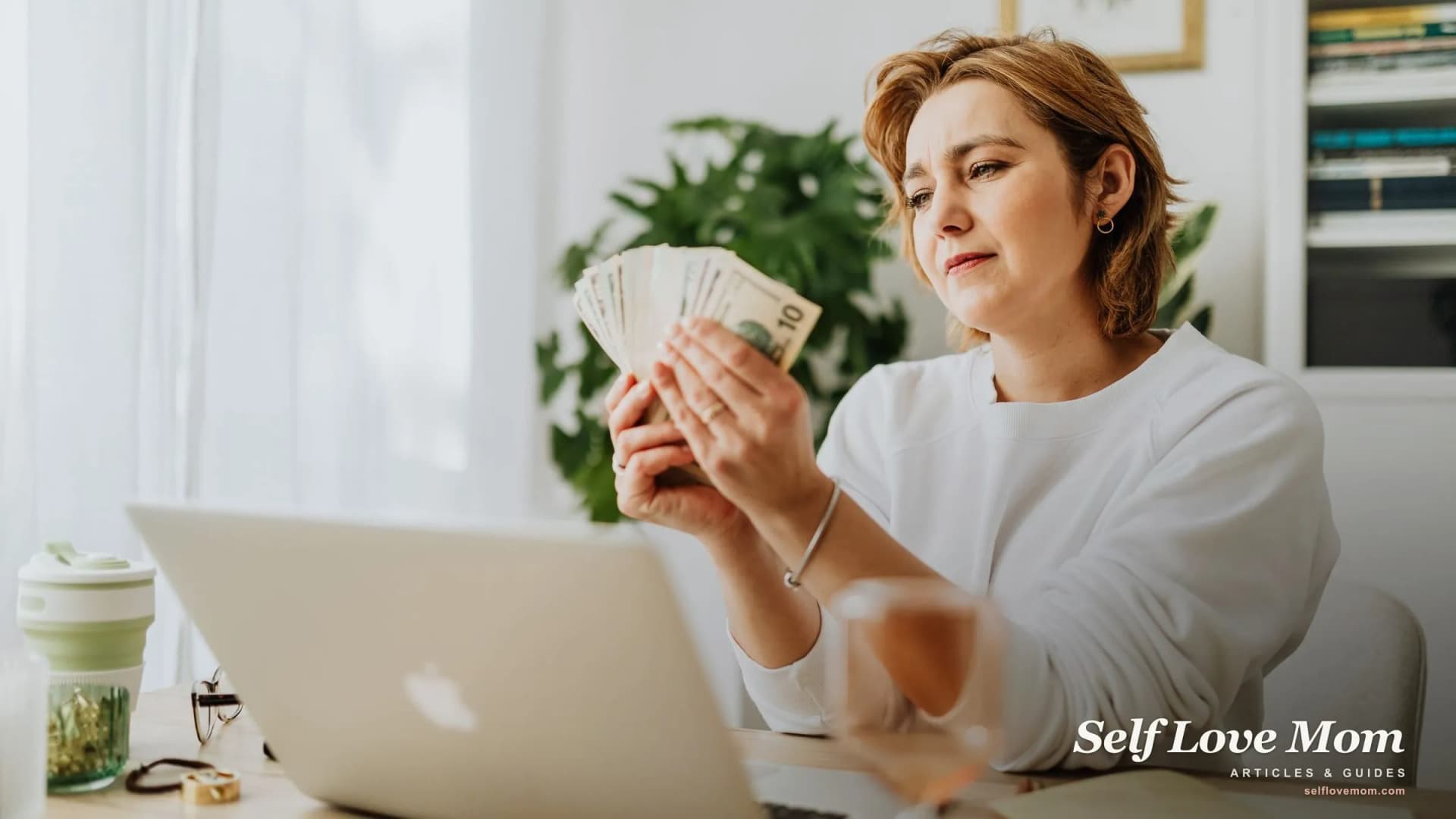 A single mom reviews cash and bills at her laptop while building a financial plan at home.