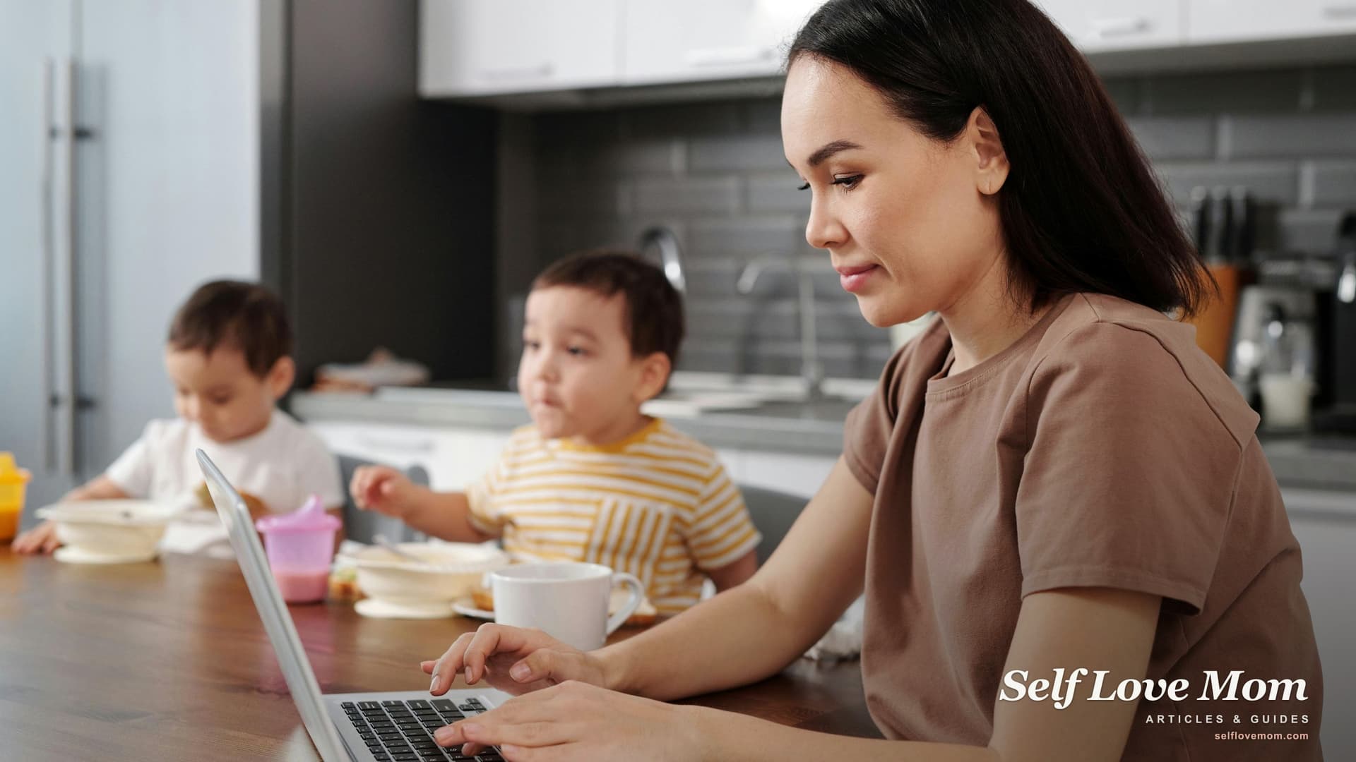 A single mom works on a laptop at the kitchen table while her two children eat breakfast, applying for help between jobs.