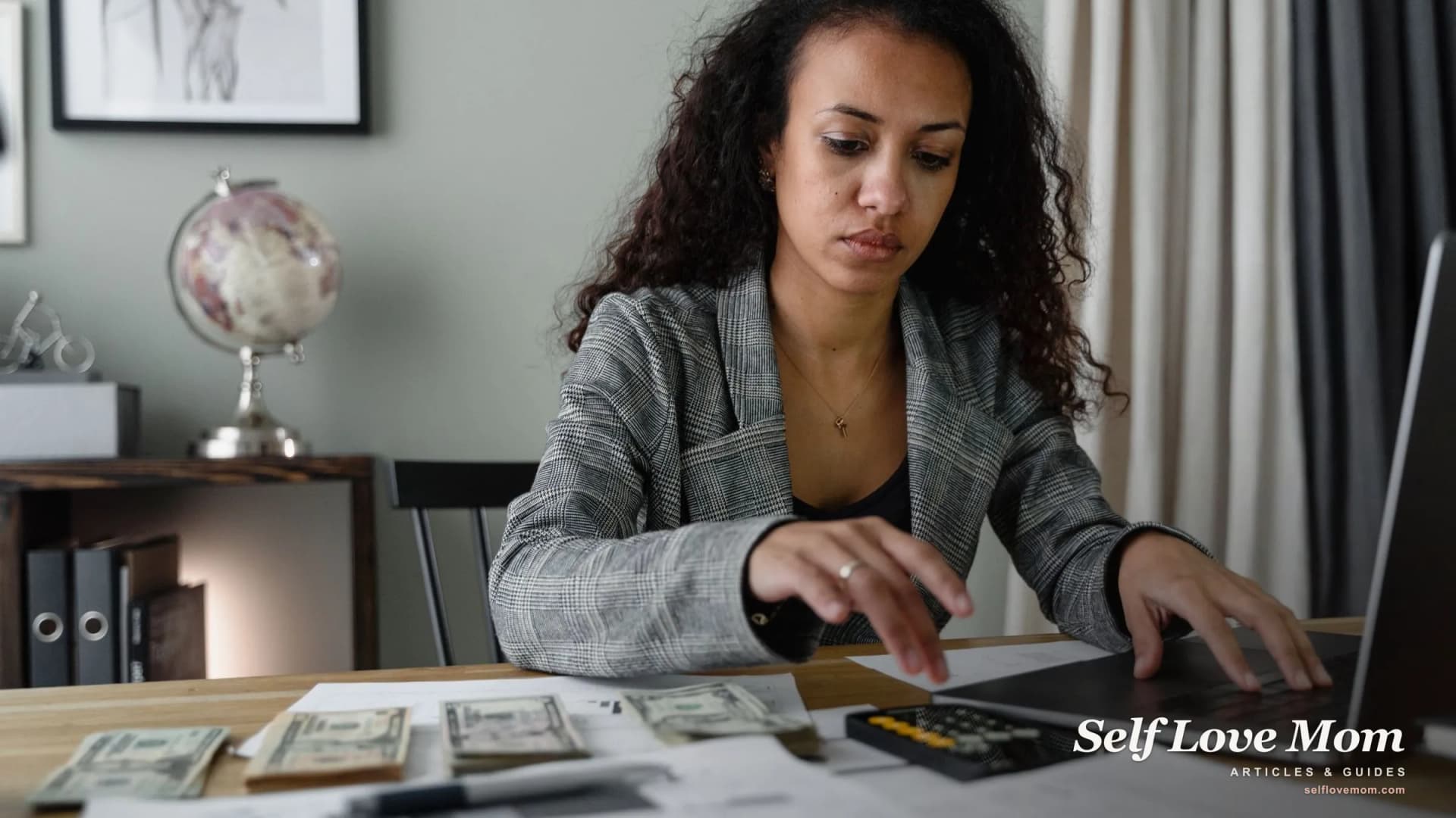 A single mom manages her income at a home desk with a laptop, calculator, and cash.