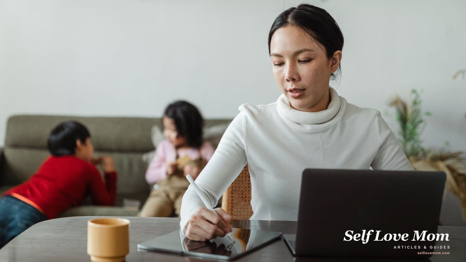A focused mother studies on a laptop and tablet at home while her two kids play behind her, the balance scholarships for working mothers help support.