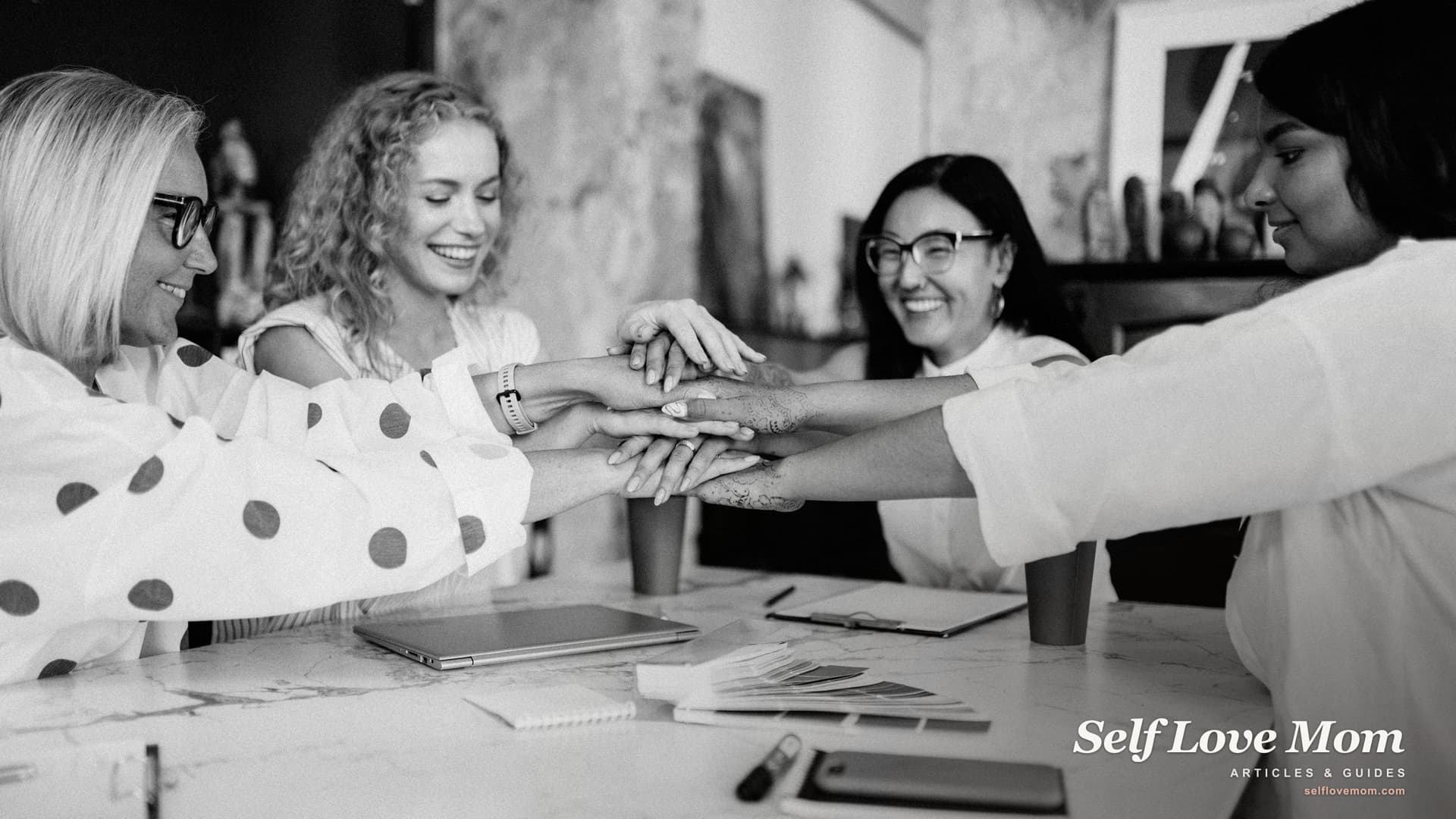 Four women put their hands together at a table, the solidarity a support group gives single moms.
