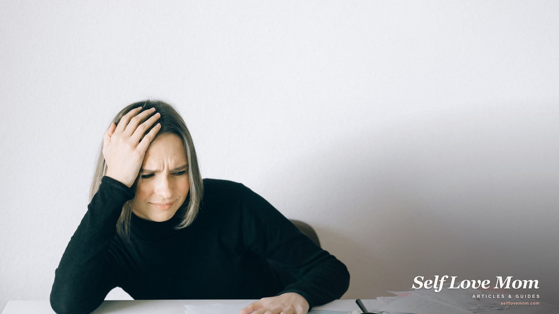 A mother alone at a table with divorce paperwork, one hand to her head, working out how to survive a divorce.