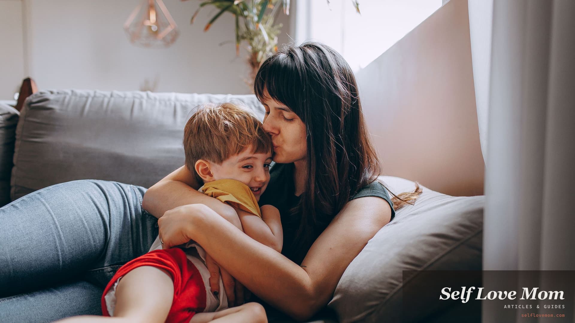 A mother kisses her young son's forehead on the couch, letting go of mom guilt after a long day.
