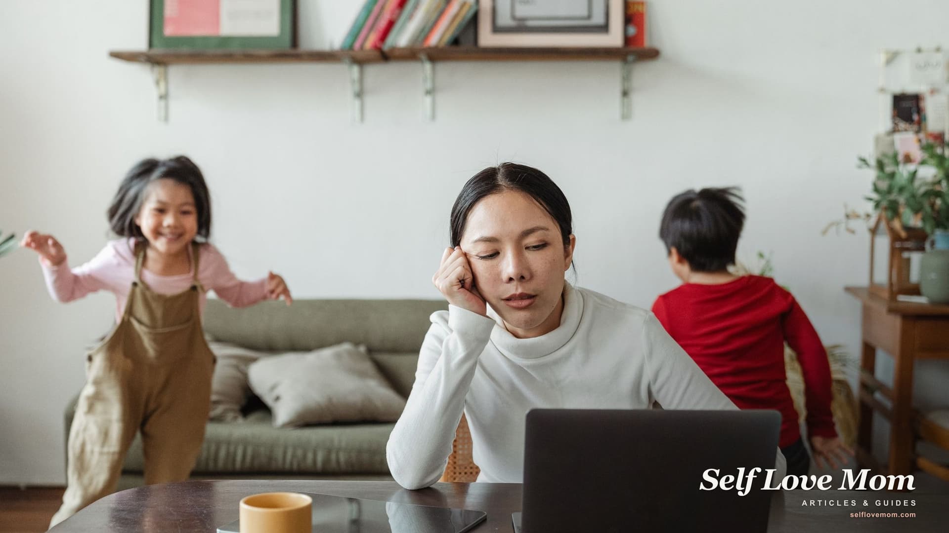 A drained mother rests her head on her hand at the kitchen table while her two children play around her.