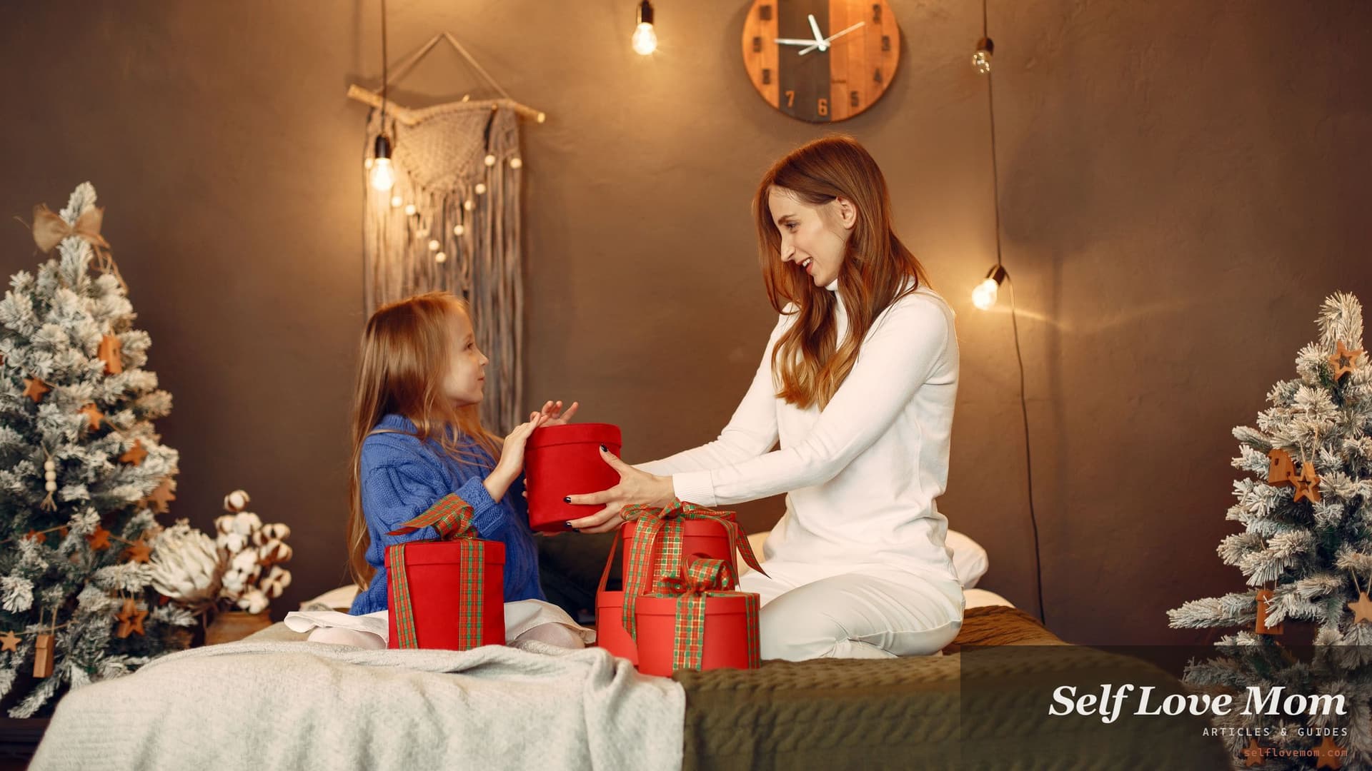 A mother hands a red wrapped gift to her young daughter as they sit together between two small Christmas trees