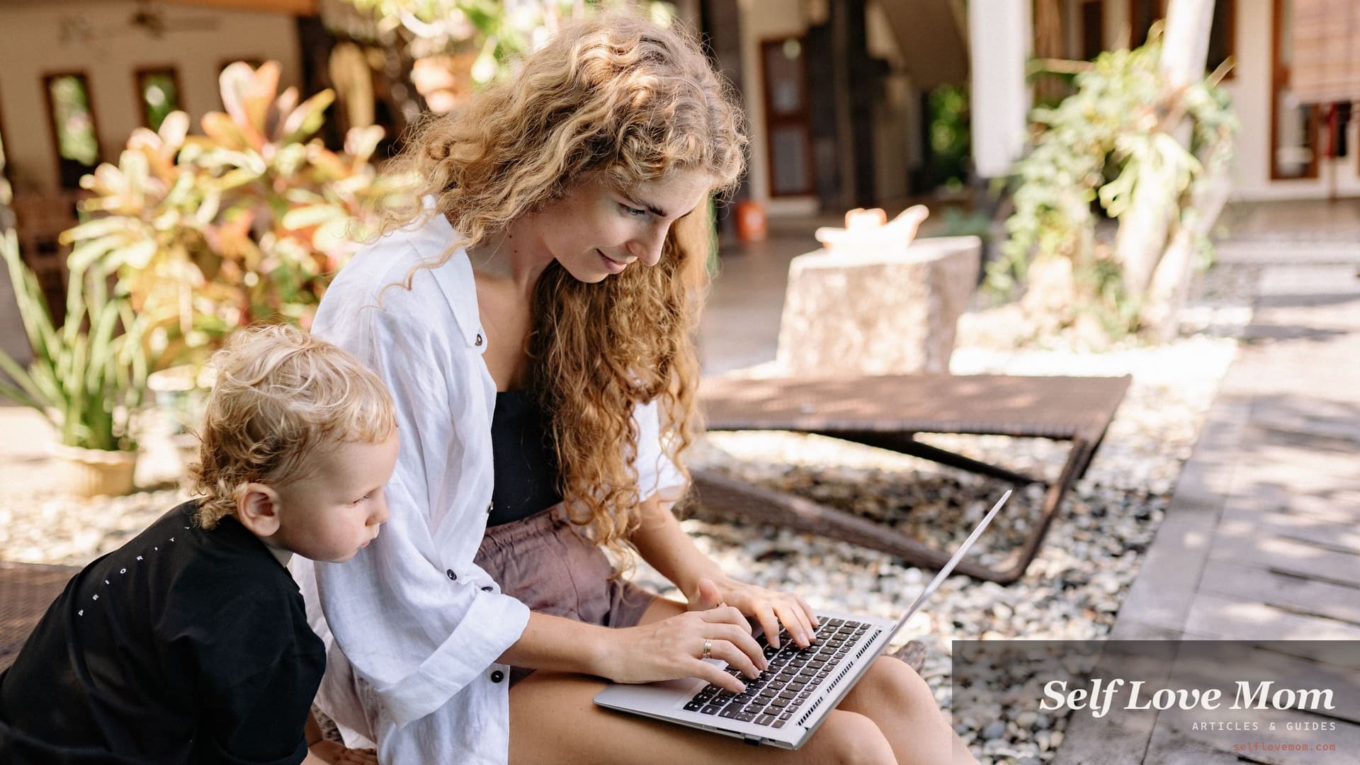 A single mom fills out grant applications on her laptop in the sunshine while her toddler leans in beside her.