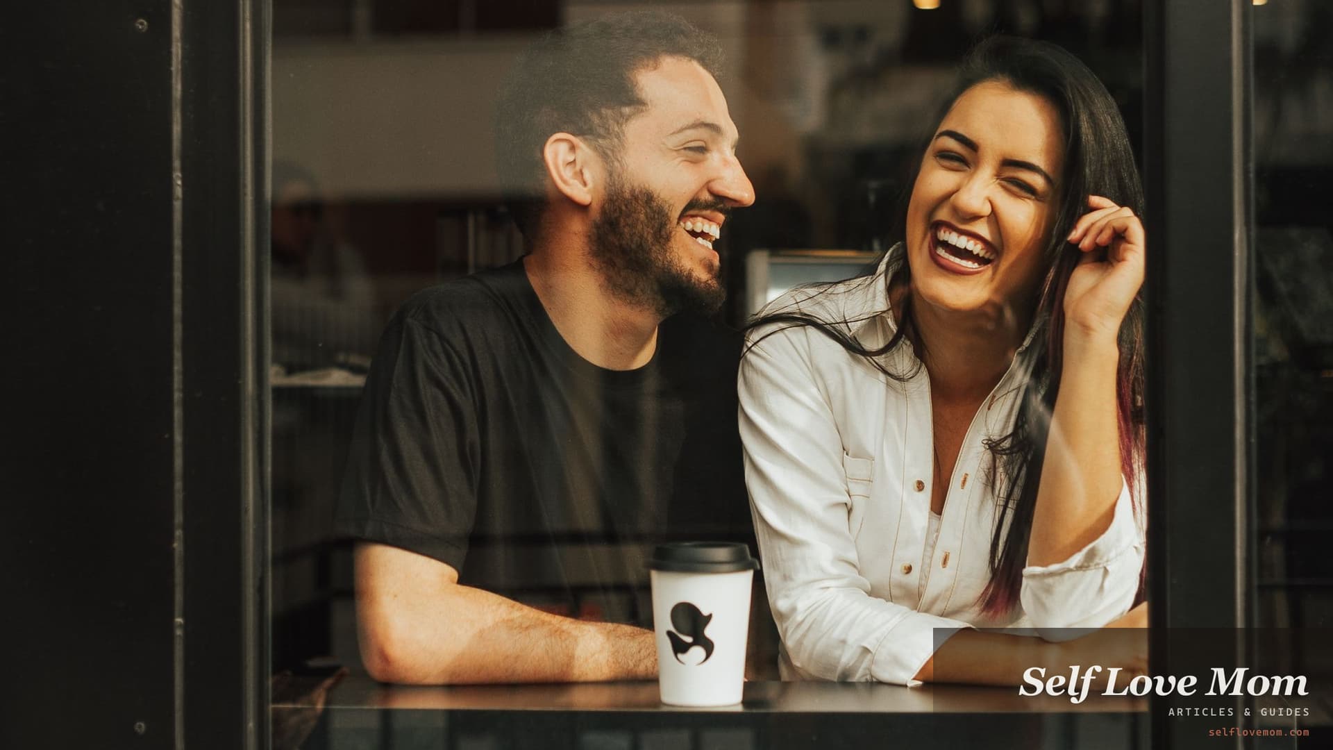 A single mom laughs over coffee on a cafe date, framed by the window at the counter.
