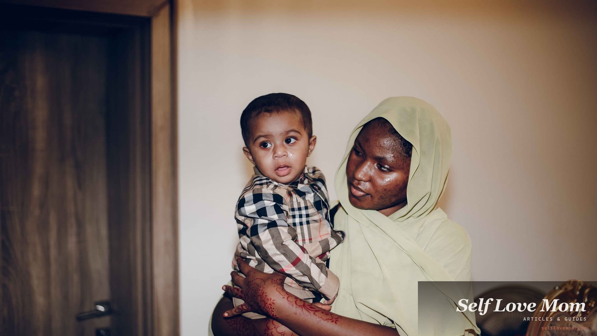 A mother in a pale green headscarf holds her toddler son on her hip in the hallway of their home, the boy looking toward the camera