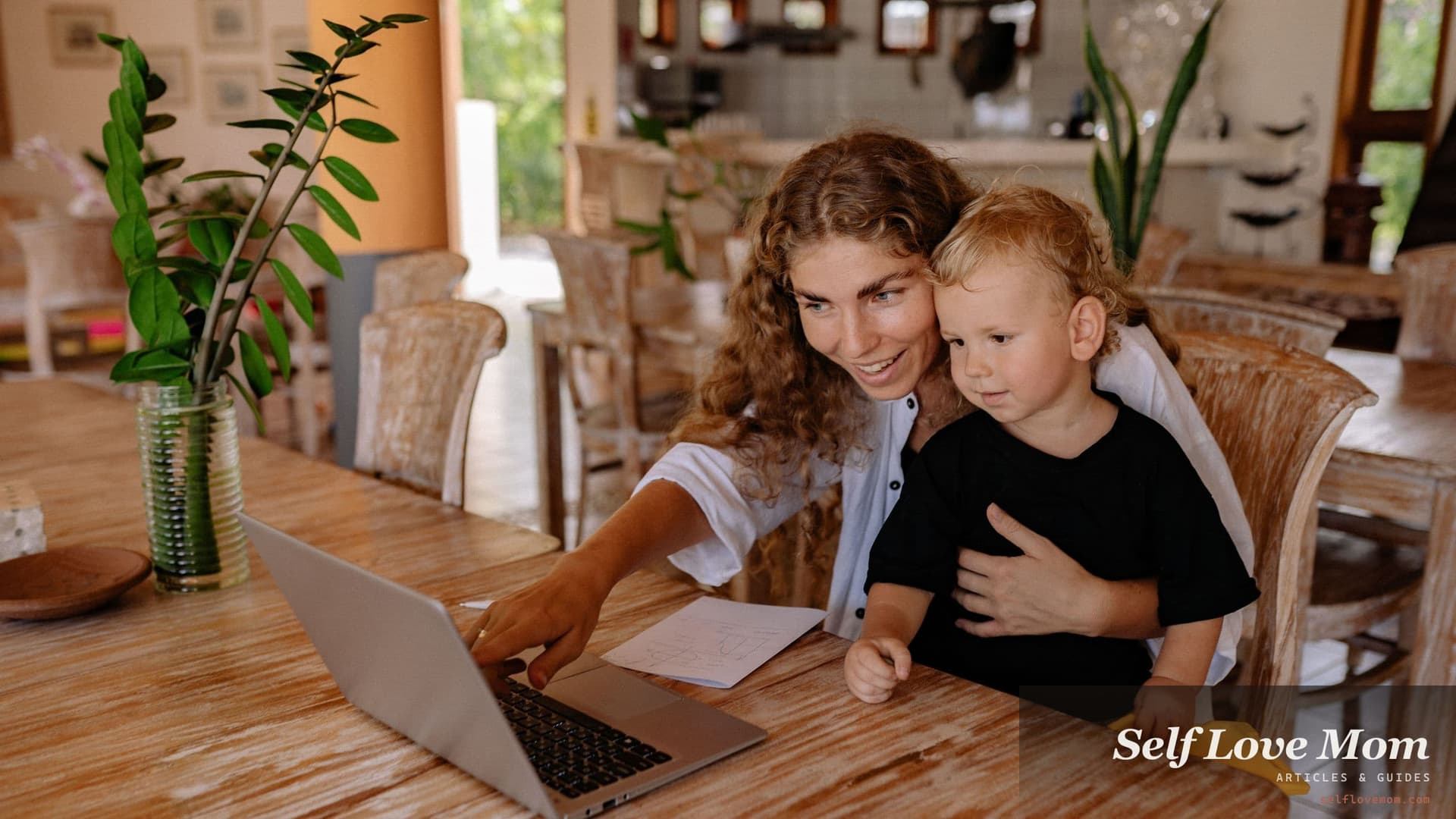 A mother points at her laptop screen with a printed form on the table beside her while her young son sits on her lap