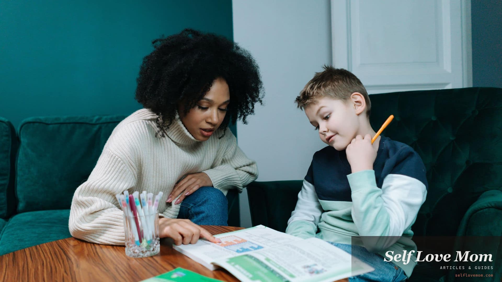 A mother sits with her young son at a table, going through a printed workbook together while he holds a pencil