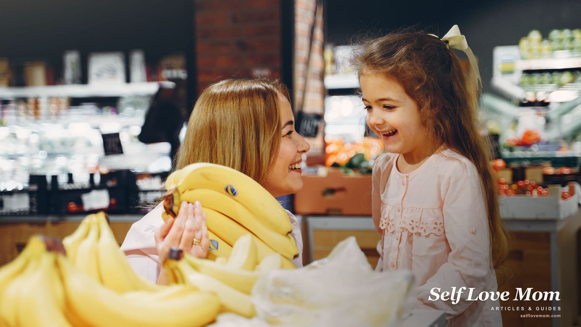 A mother and her young daughter laugh over a bunch of bananas in a grocery store produce aisle, the everyday shopping trip that EBT discounts stretch further.