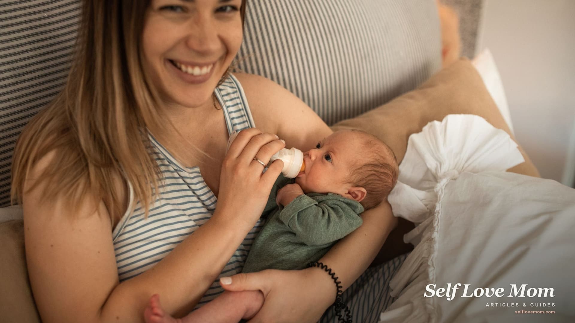 A young mother smiles at the camera while bottle-feeding her newborn baby at home.