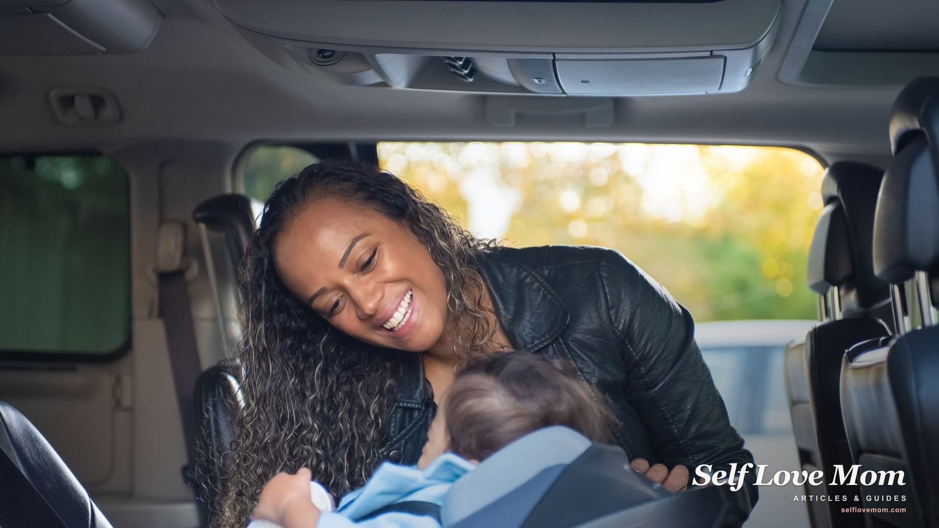 A smiling mother leans into the back seat of a car to check on her baby, who is buckled into an infant car seat.