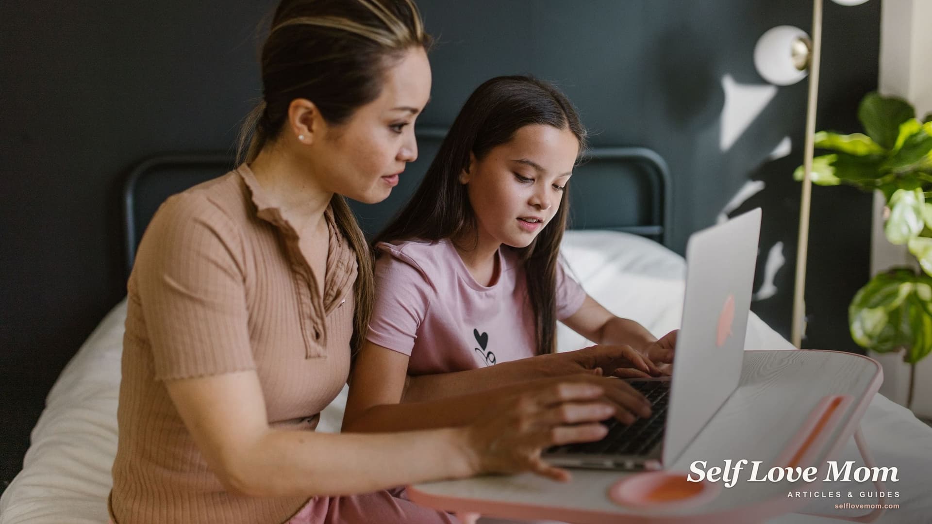 A mother sits beside her daughter on the bed and guides her hands on a laptop keyboard, the kind of refurbished machine the free laptop programs deliver.