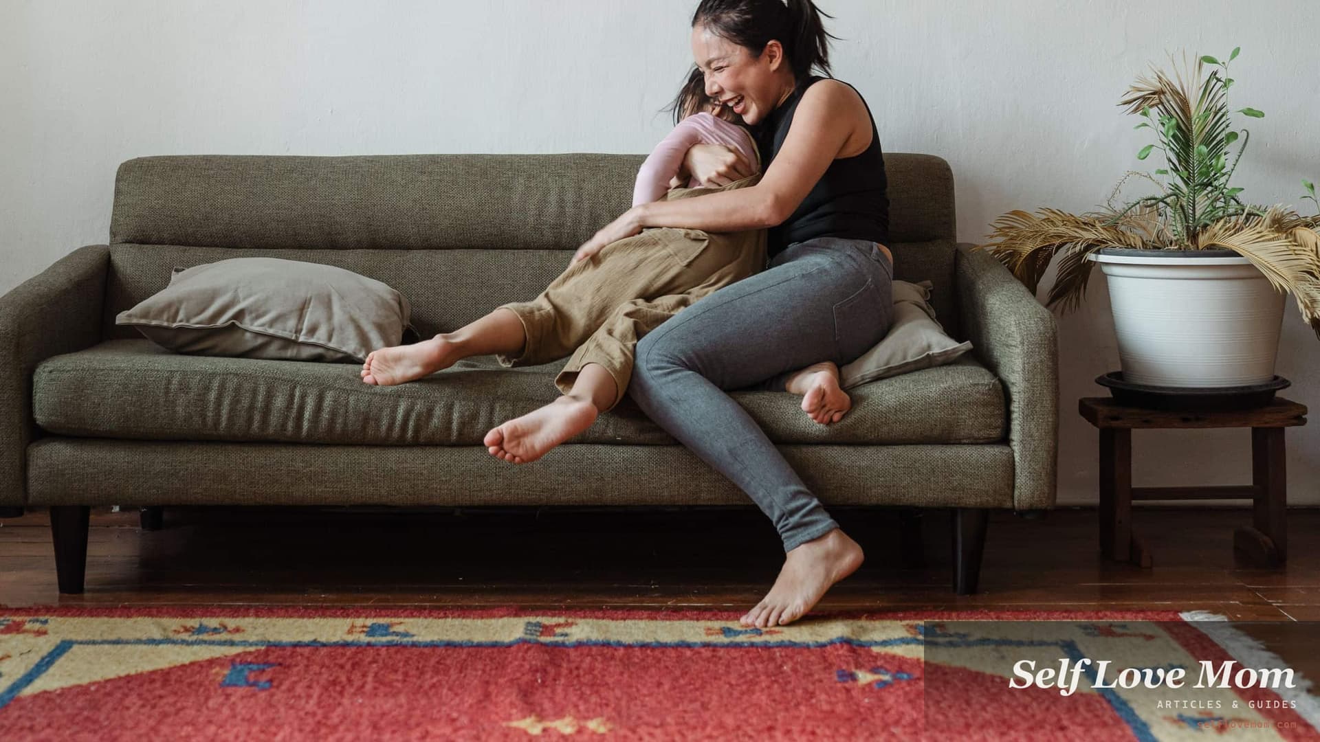 A single mother laughs with her young daughter on the sofa of their apartment.