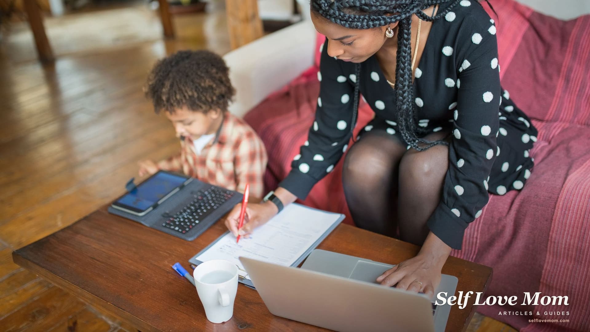 A mother fills in a paper application at a coffee table with a laptop open while her young son plays on a tablet beside her.