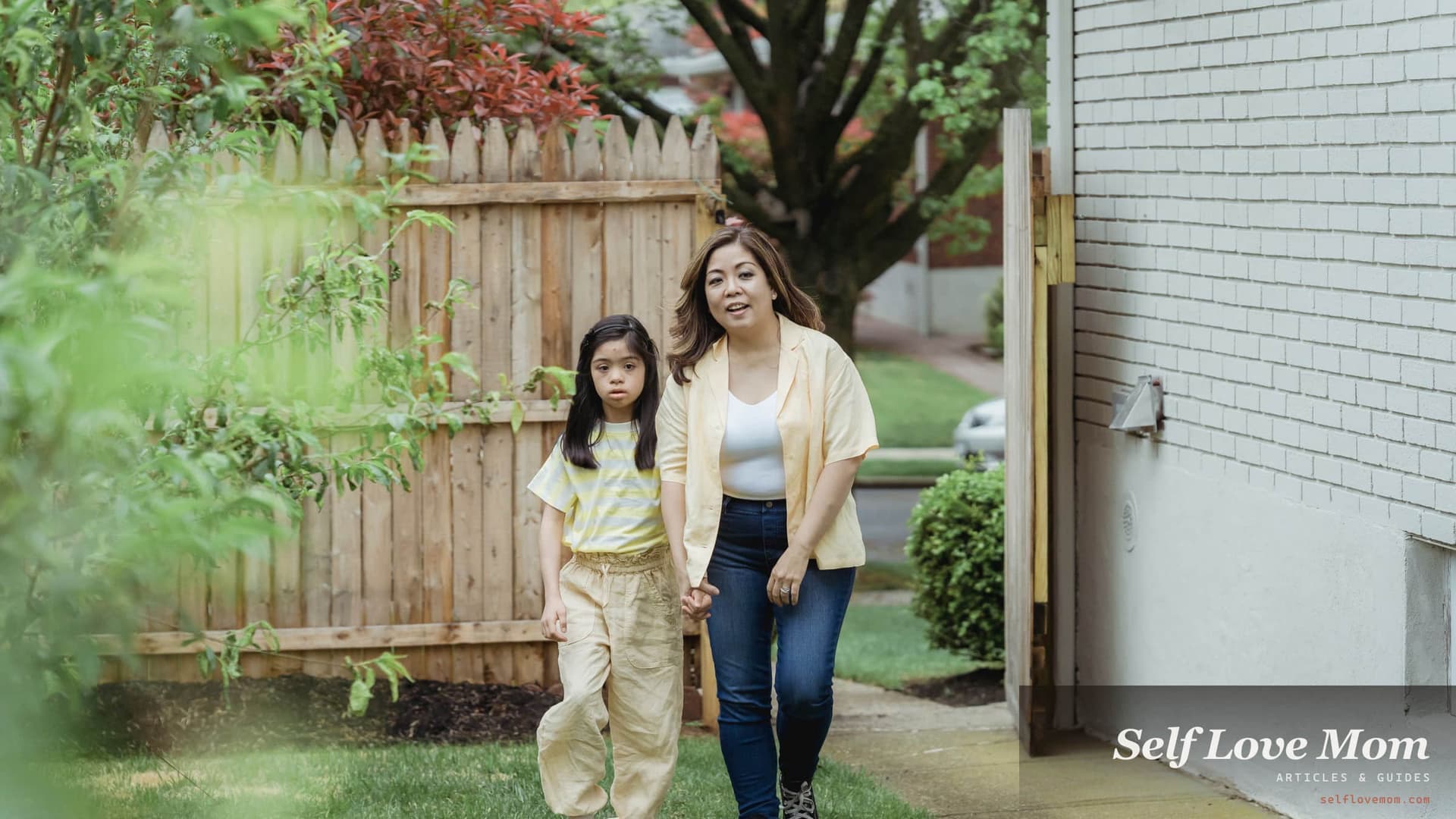 A single mother and her daughter walk hand in hand up the path to their home past a wooden fence.