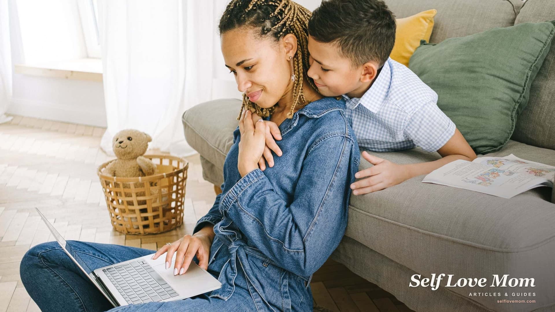A single mom studies on her laptop on the living room floor while her young son leans over the couch to hug her.
