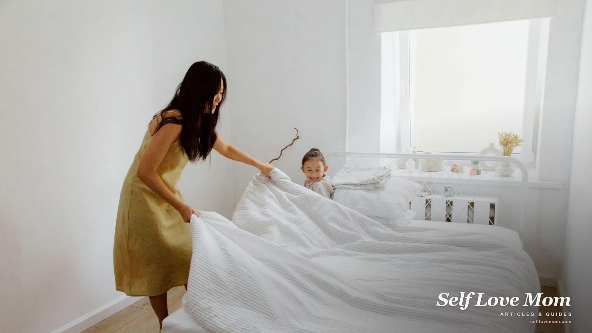 A mother spreads a clean white duvet over her daughter's new bed in a bright bedroom while the little girl watches from the pillows.
