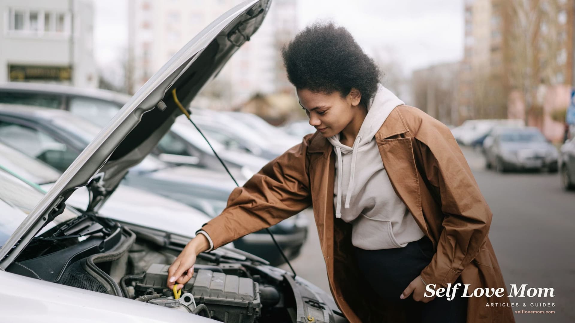 A young woman in a brown coat checks the engine oil under the open hood of her car on a city street.
