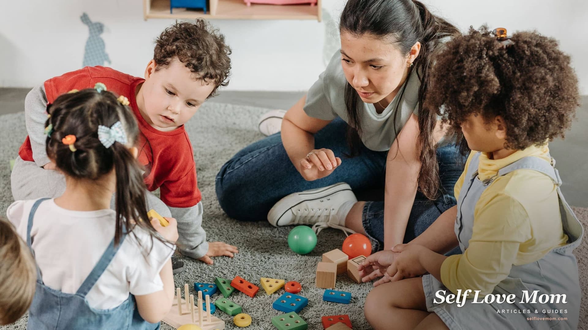 A child care teacher sits on the classroom floor with four toddlers, helping them play with stacking toys, the everyday work a CDA credential prepares you for.