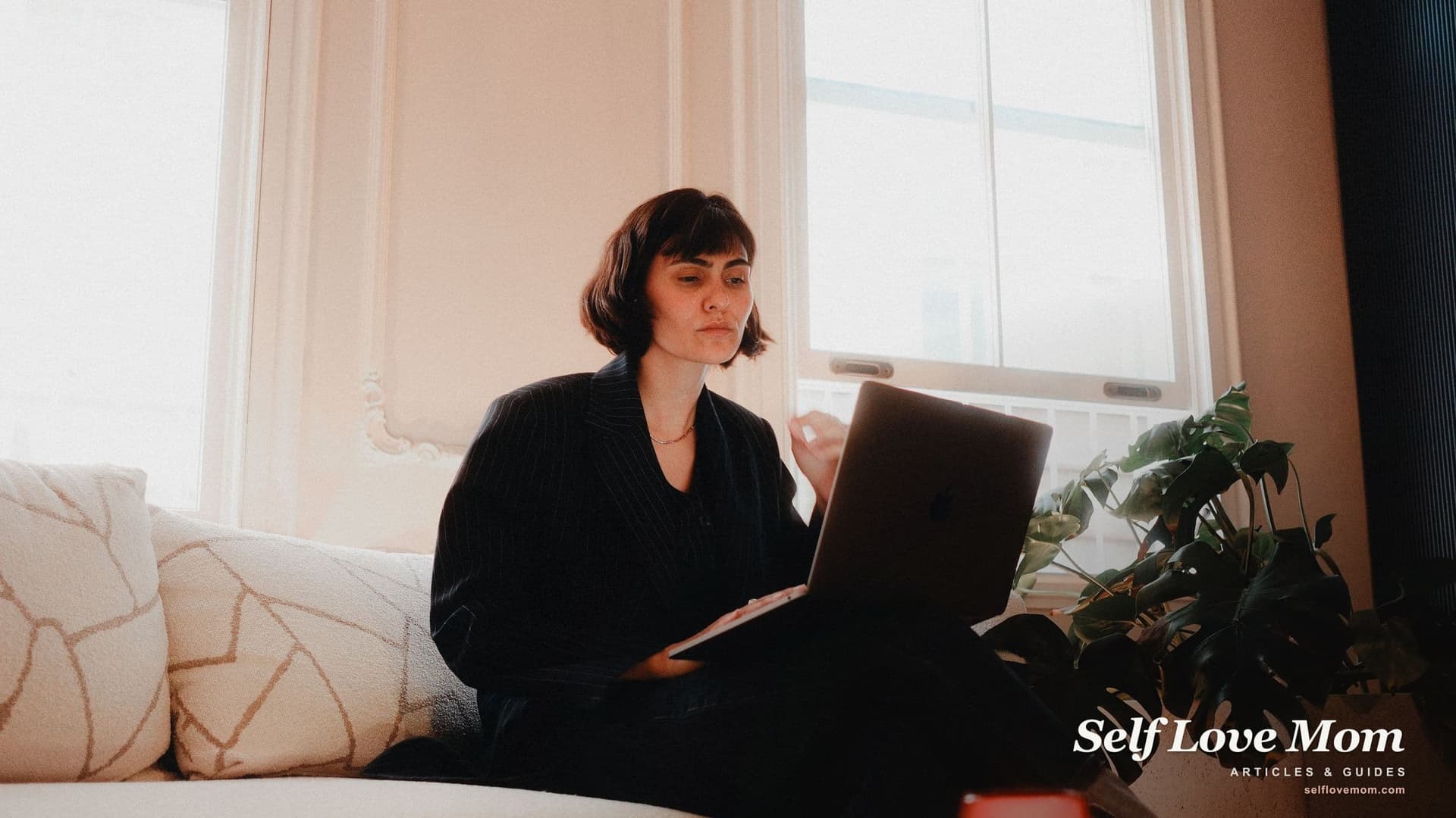 A woman sits on a sofa by a sunny window, working on a laptop on her lap.