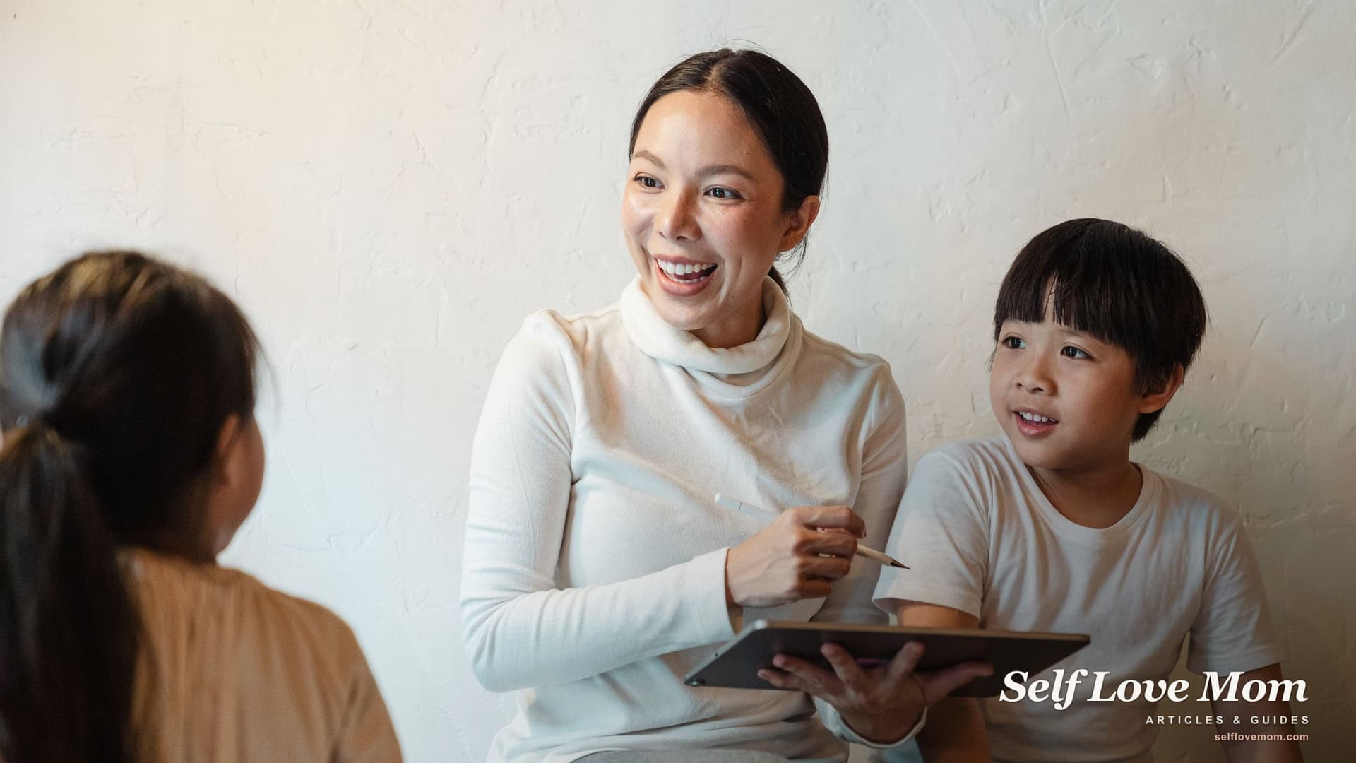 A smiling mother holds a tablet and a stylus as her young son and daughter sit with her in a bright living room.