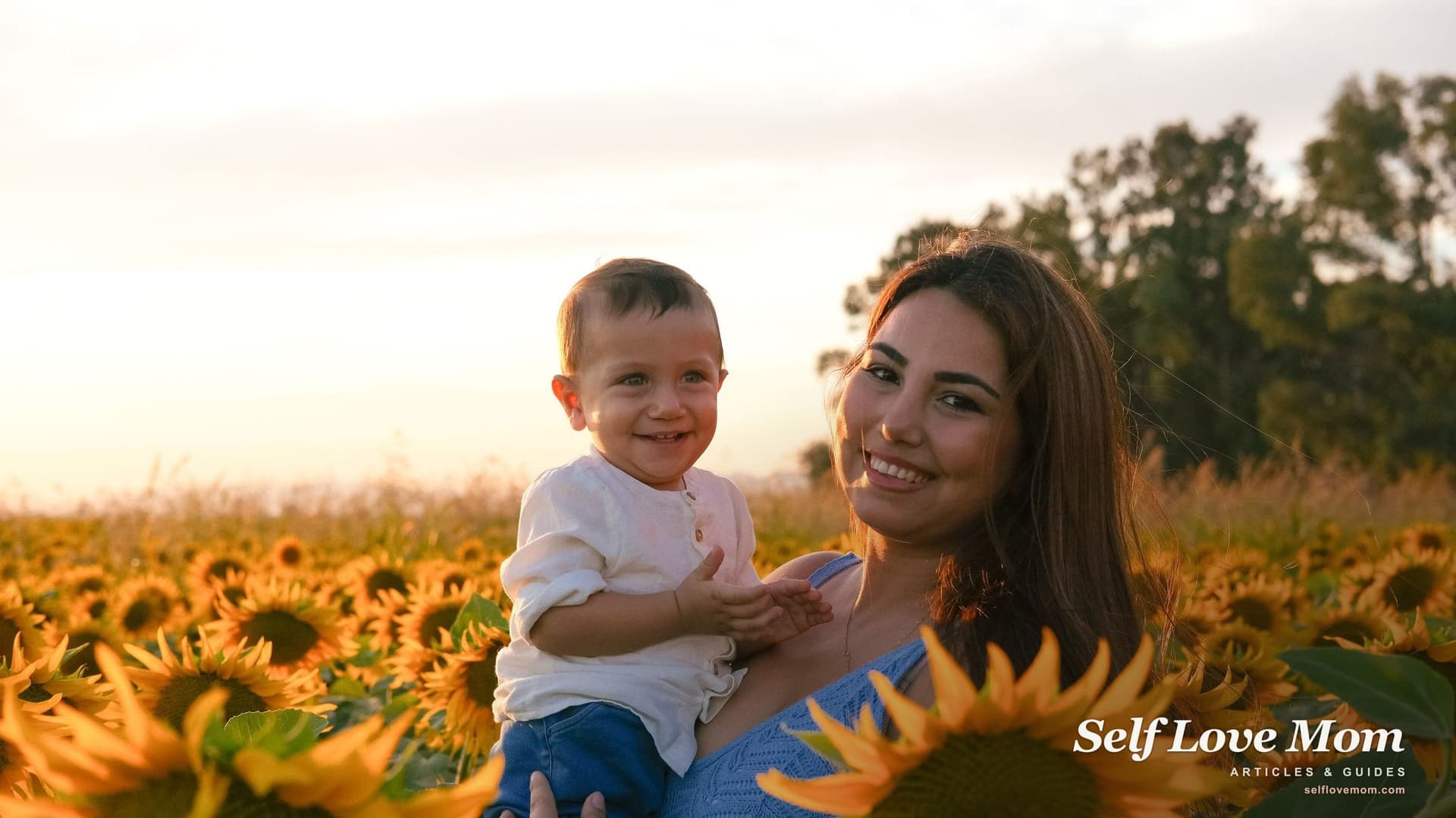 A smiling single mom holds her toddler son in a sunflower field at sunset, the kind of family that grants for single mothers in Kansas are built to support.