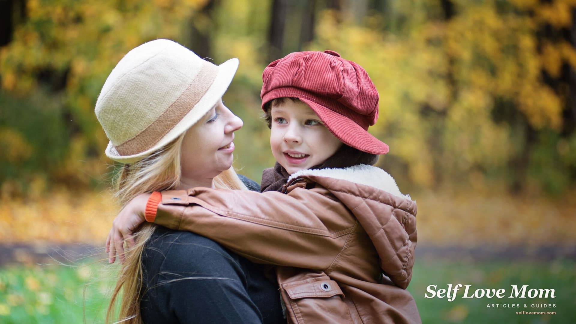 A smiling mother in a cream sun hat holds her young child, who wears a red cap and a brown jacket, among autumn trees.